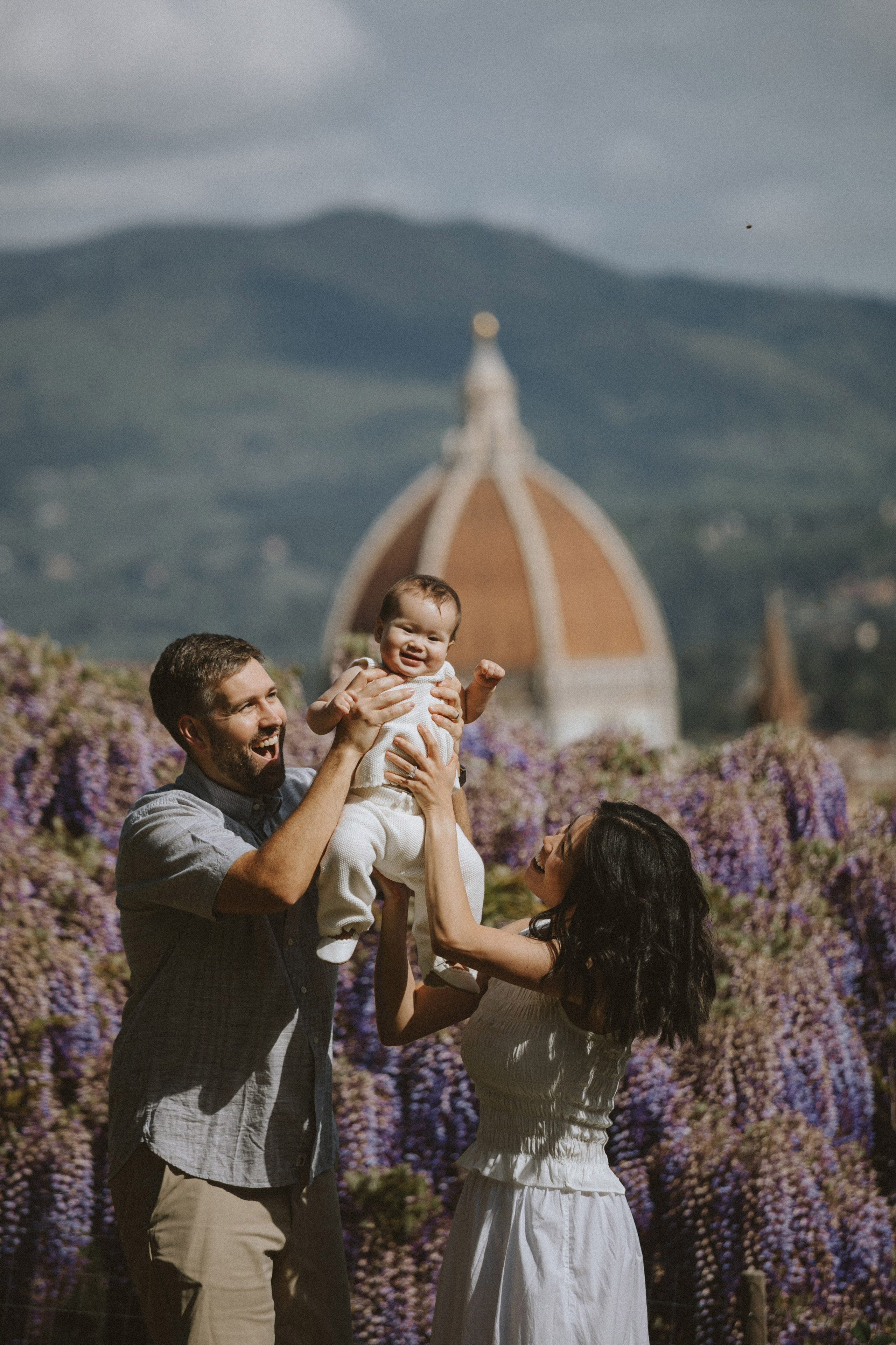 Genitori felici che sollevano la figlia piccola con la Cupola del Brunelleschi e i giardini di glicine sullo sfondo.