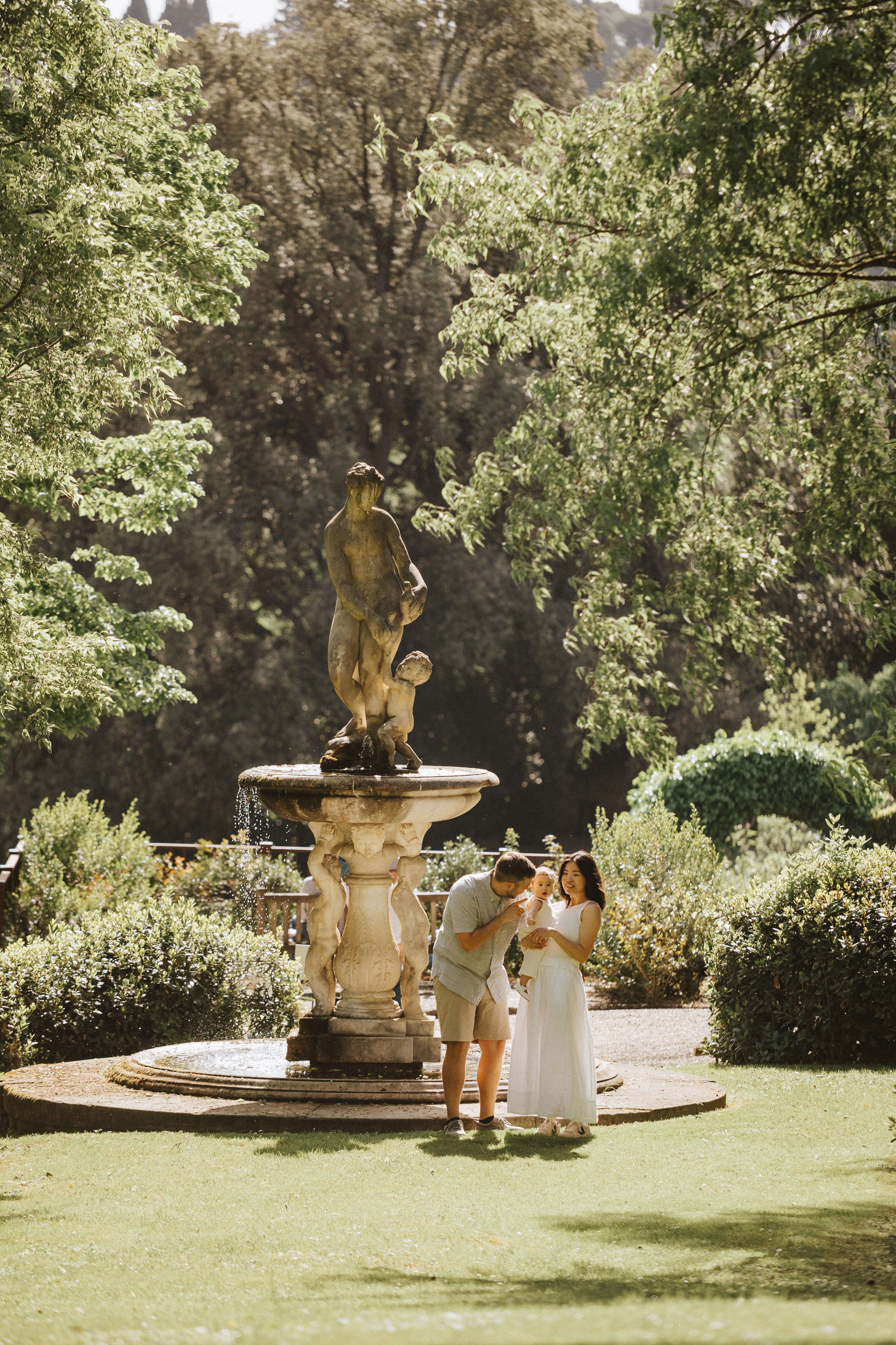 Famiglia in posa accanto a una storica fontana in pietra nei rigogliosi giardini di Villa Bardini in Toscana.