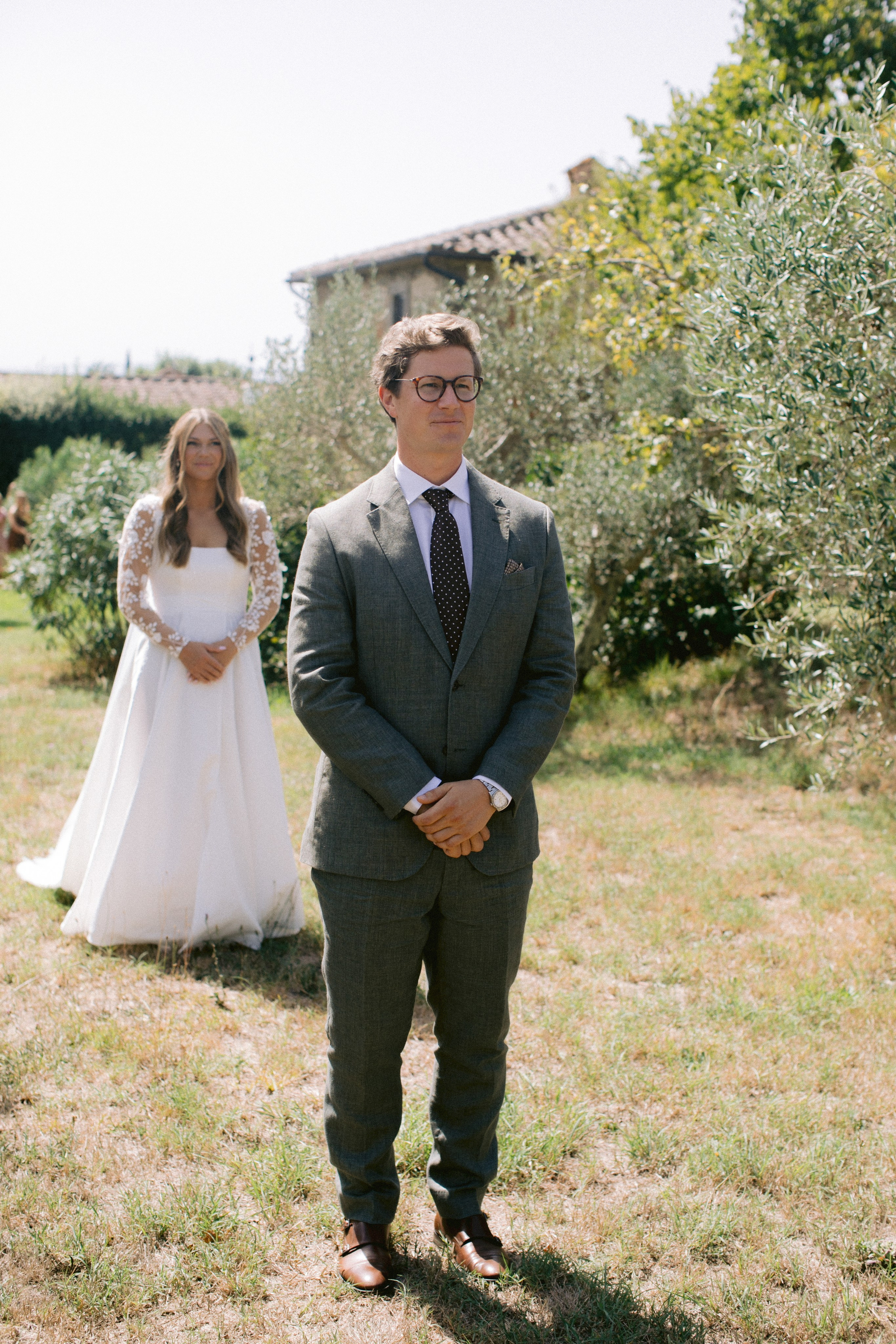 "Primer vistazo de los novios en un pintoresco jardín en Italia: el novio con un elegante traje gris está al frente, mientras que la novia con un vestido blanco elegante y mangas de encaje está detrás de él."