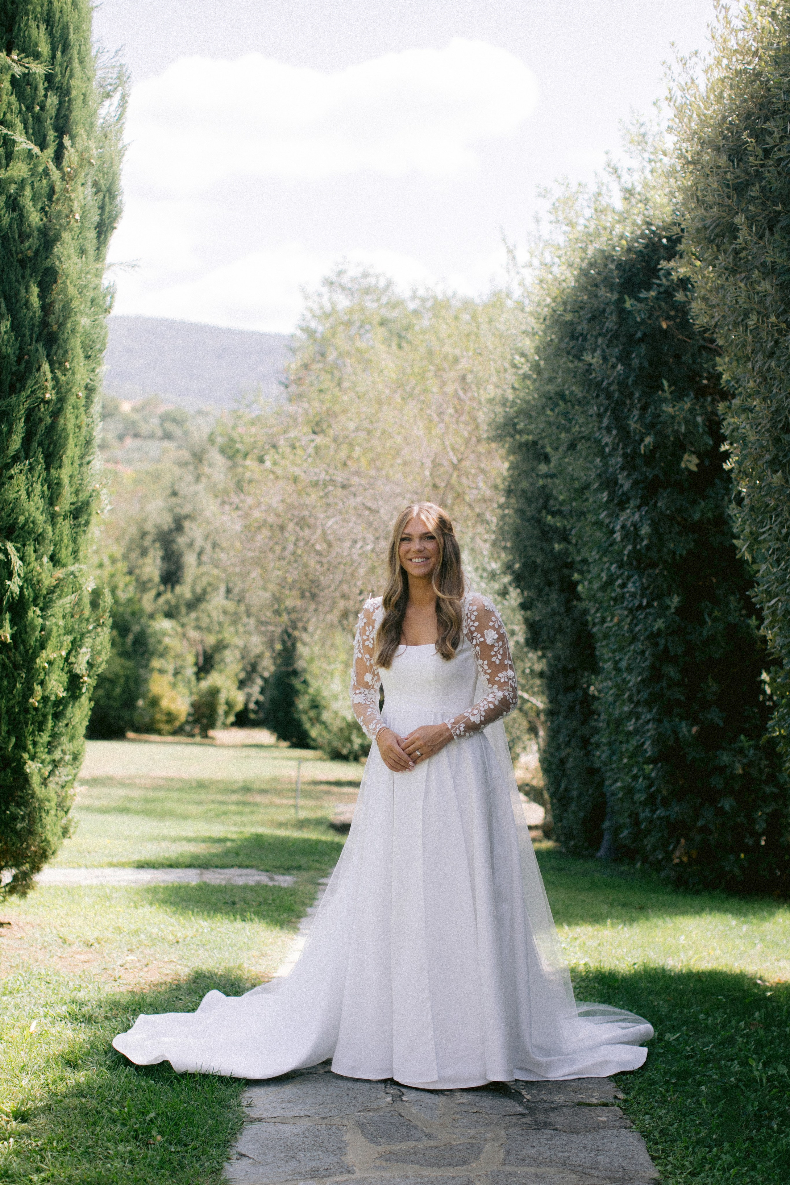 "Novia con un elegante vestido blanco de mangas de encaje, de pie en un pintoresco jardín en Cortona, Italia, rodeada de cipreses verdes y colinas escénicas."