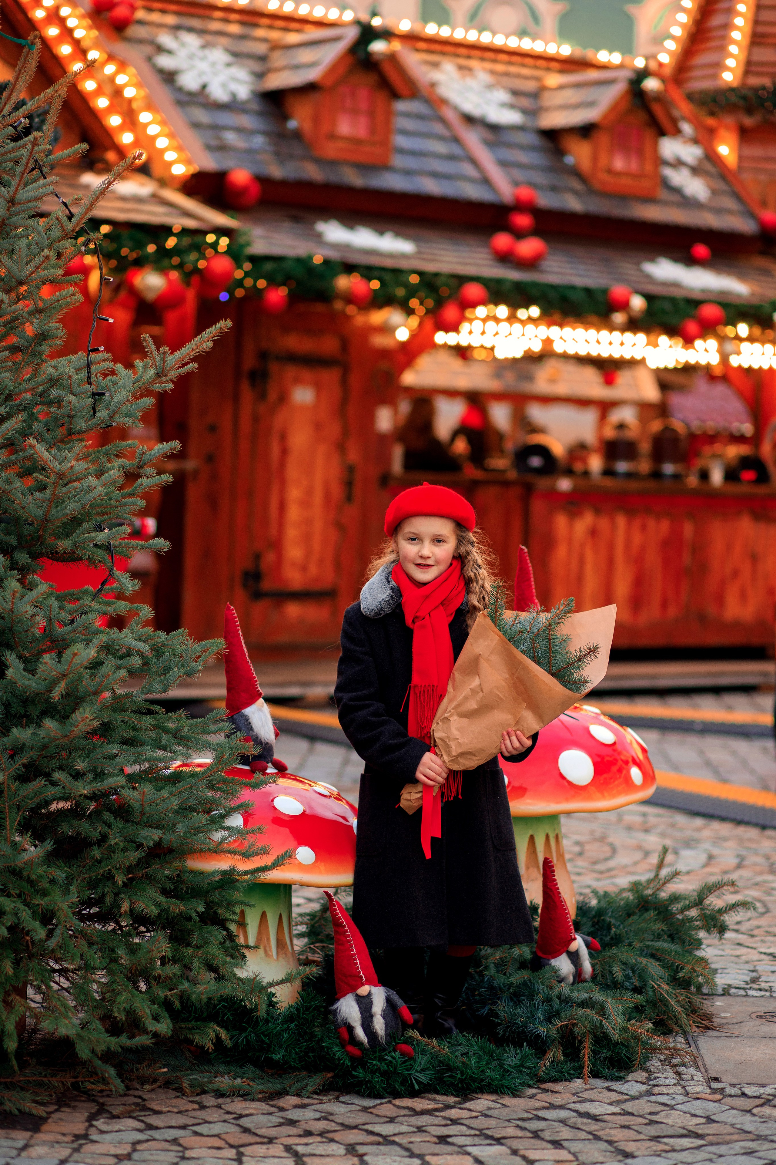 Дівчинка тримає букет з гілок, поряд з нею ялинка і мухомори, а позаду прилавок з вогниками