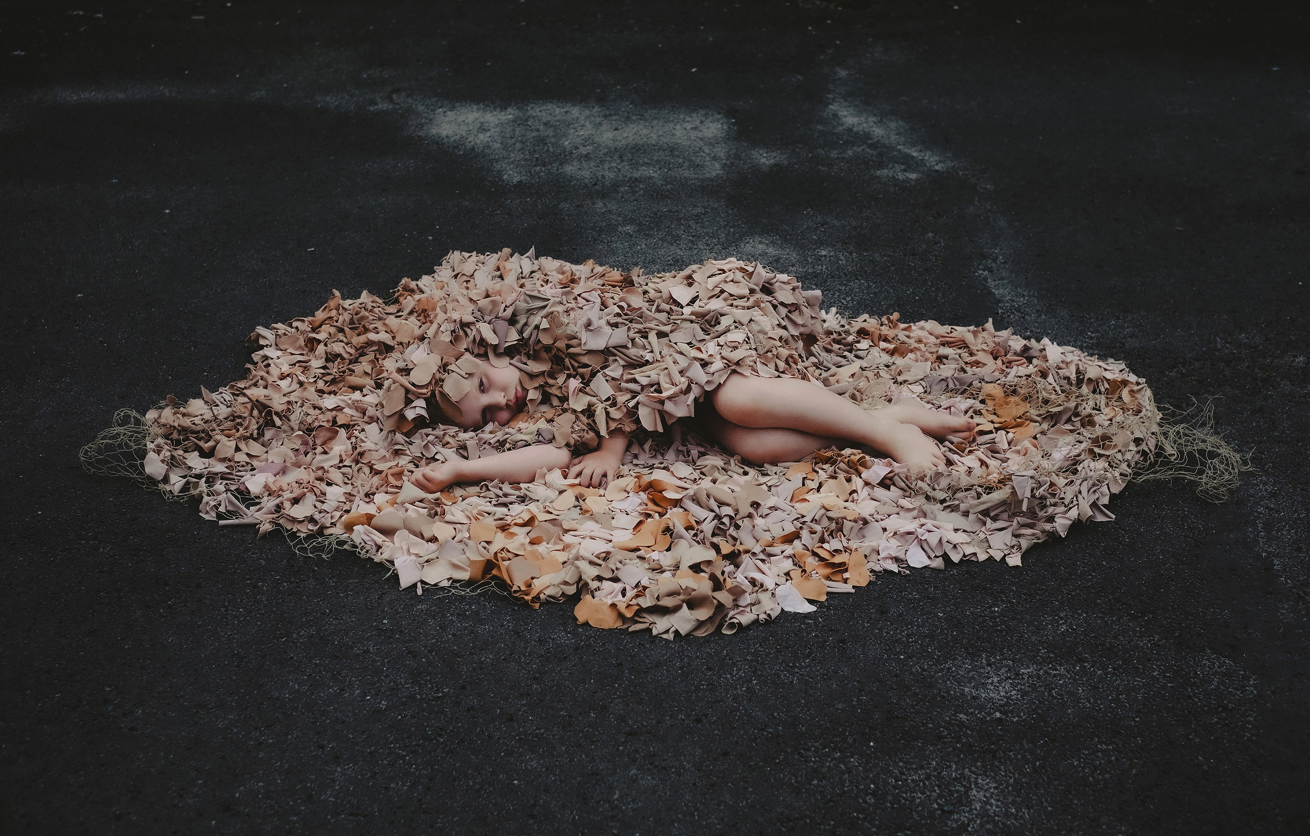 A young boy is lying on the ground, covered by a large net made of materials in various shades of beige and brown, resembling human skin tones. The net, part of the "Azyl" (Asylum) project, envelopes his body almost entirely, symbolizing protection and vulnerability. He appears peaceful and serene, contrasting with the dark, rough texture of the ground beneath her. This artwork conveys themes of refuge, intimacy, and the human condition in the context of conflict and displacement, highlighting the project’s focus on providing a metaphorical shelter from violence and war.