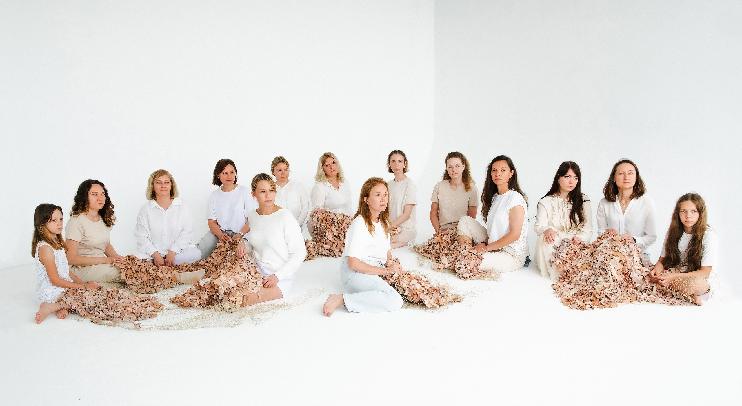 A group of women and girls from various backgrounds are sitting together on the floor in a bright, white room. They are working on weaving a large net made of materials in shades resembling human skin tones. The participants, dressed in light and neutral-colored clothing, include individuals of different ages. This collaborative art project, titled "Azyl" (Asylum), serves as a metaphor for a shelter for humanity, providing refuge from violence and war. The project highlights the legal and social issues faced by women, especially in the context of political and military tensions, and emphasizes the role of women in conflict prevention, resolution, and the use of feminist activism.
