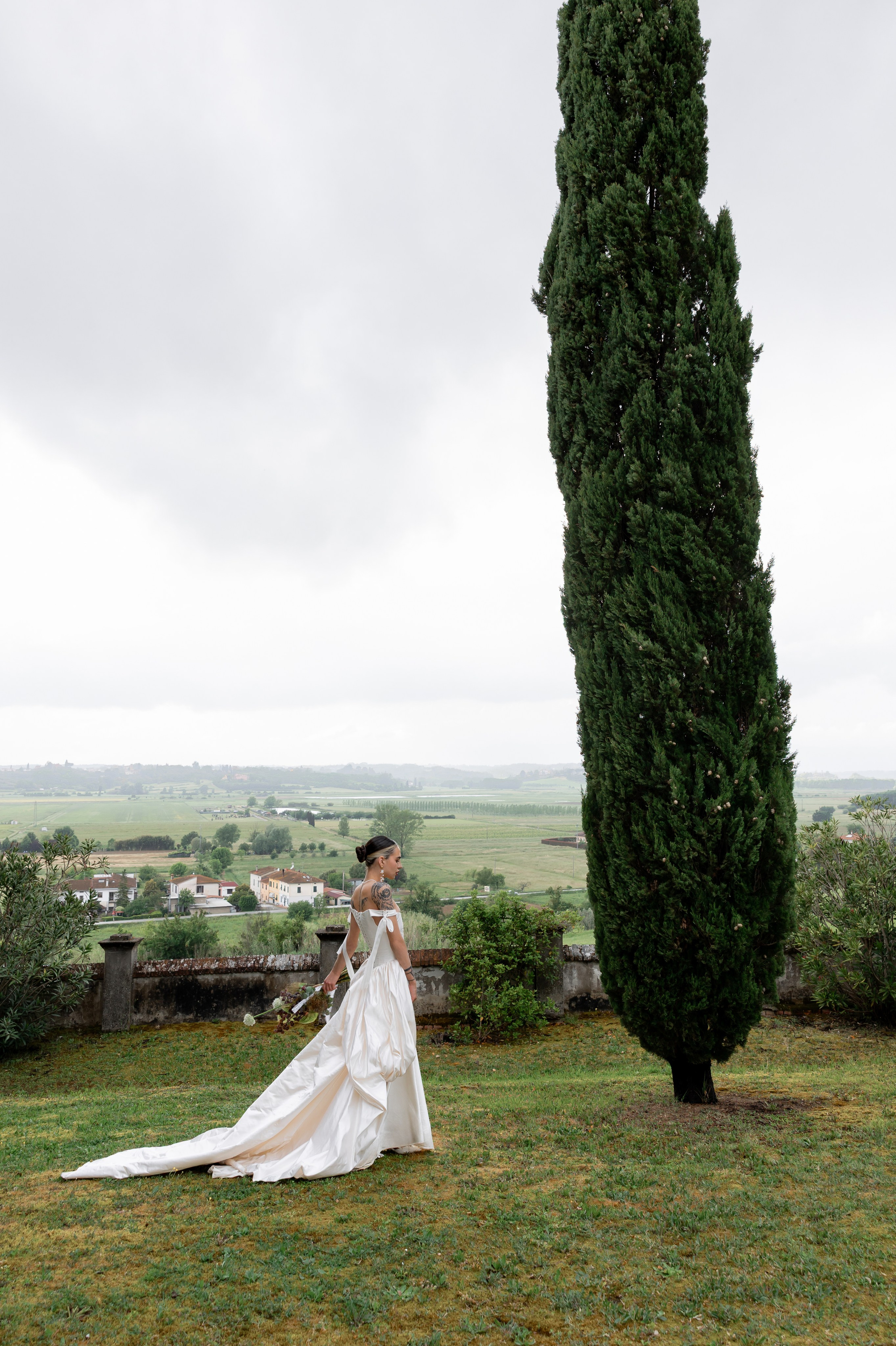 Bride walking through the Tuscany countryside surrounded by cypress trees and dramatic cloudy sky.