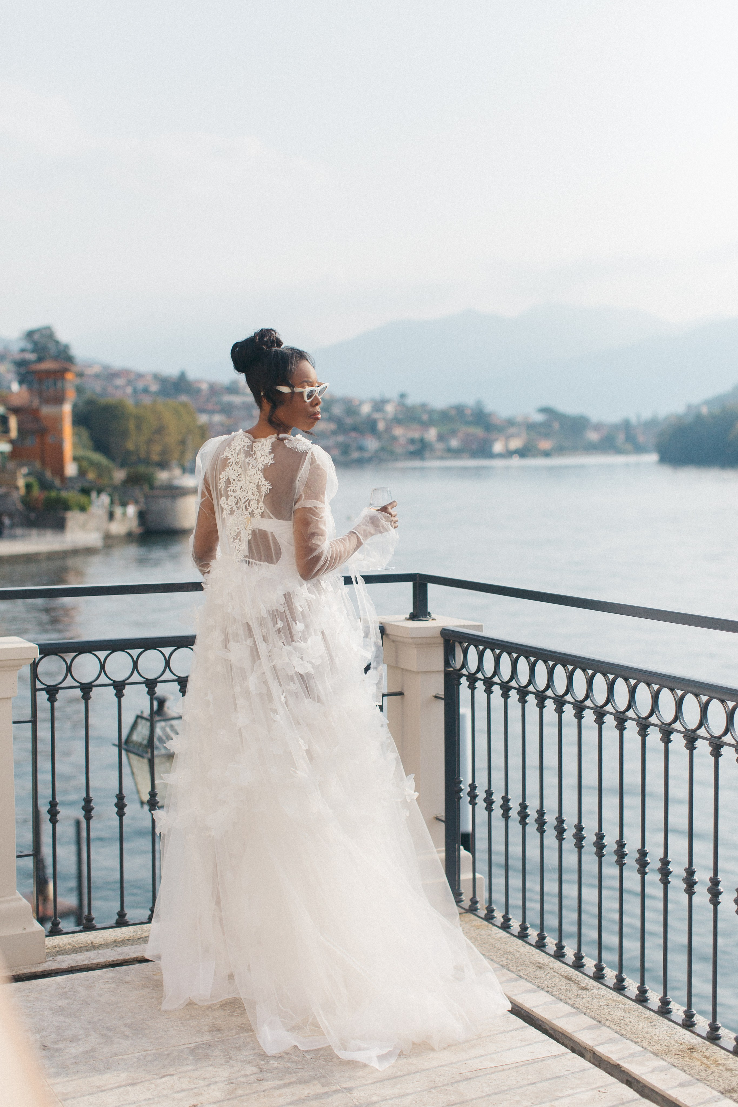 Bride in silk robe on balcony, holding wine and looking out over Lake Como