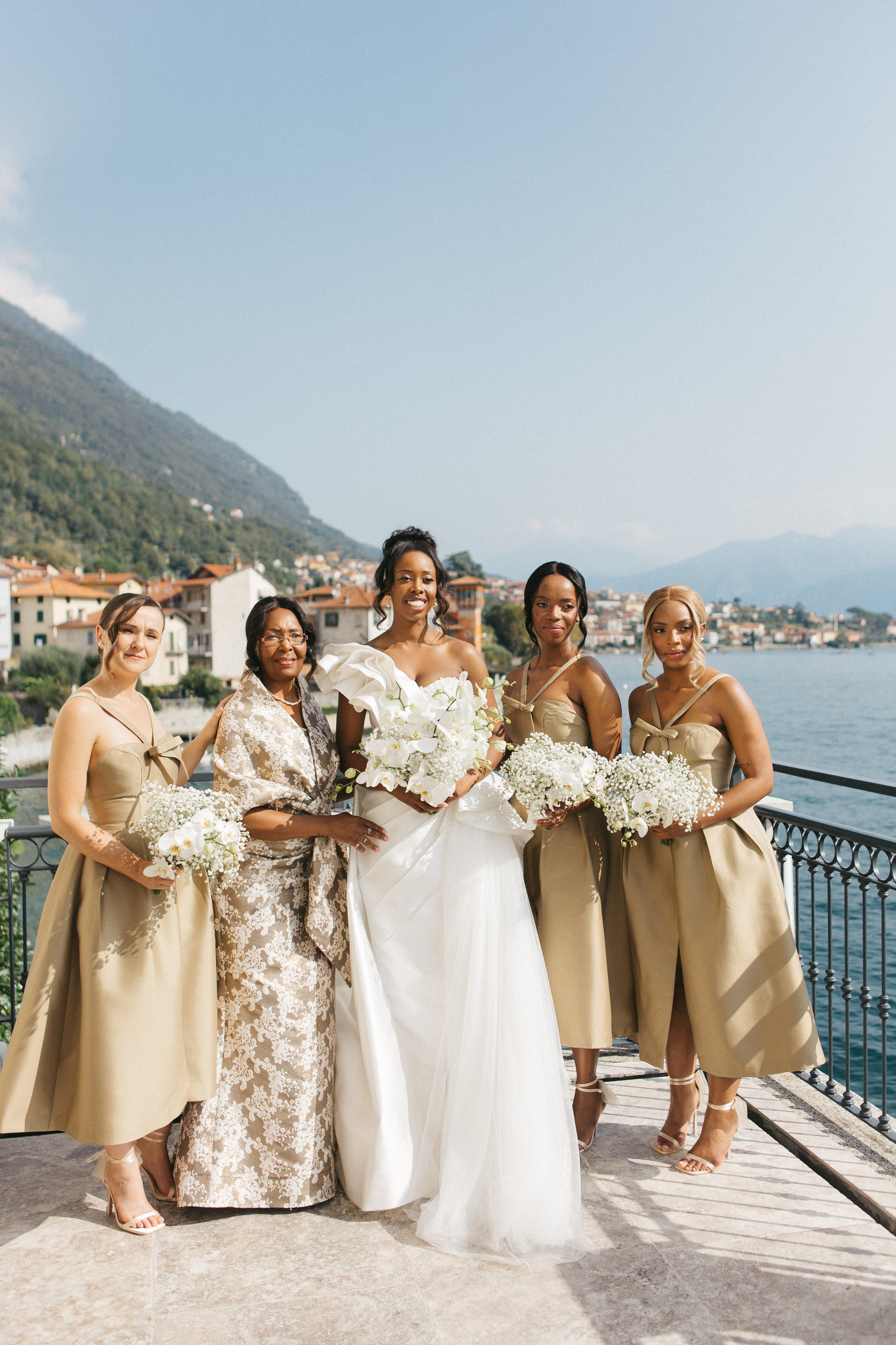 Elegant portrait of a dark-skinned bride with her bridesmaids on a balcony, scenic Lake Como in the background in bright classical wedding photography style 
