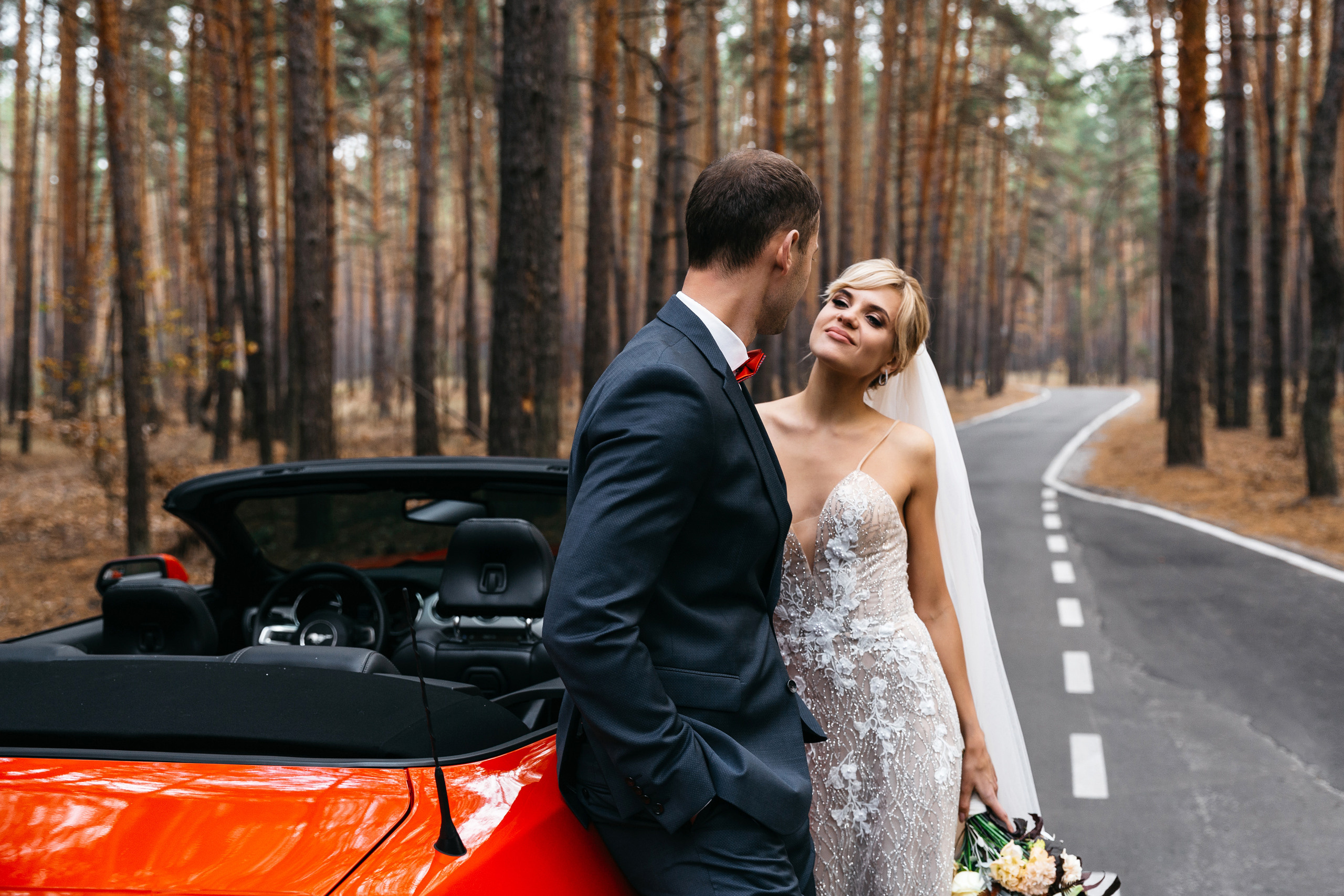 жених и невеста позируют в сосновом лесу с красным кабриолетом