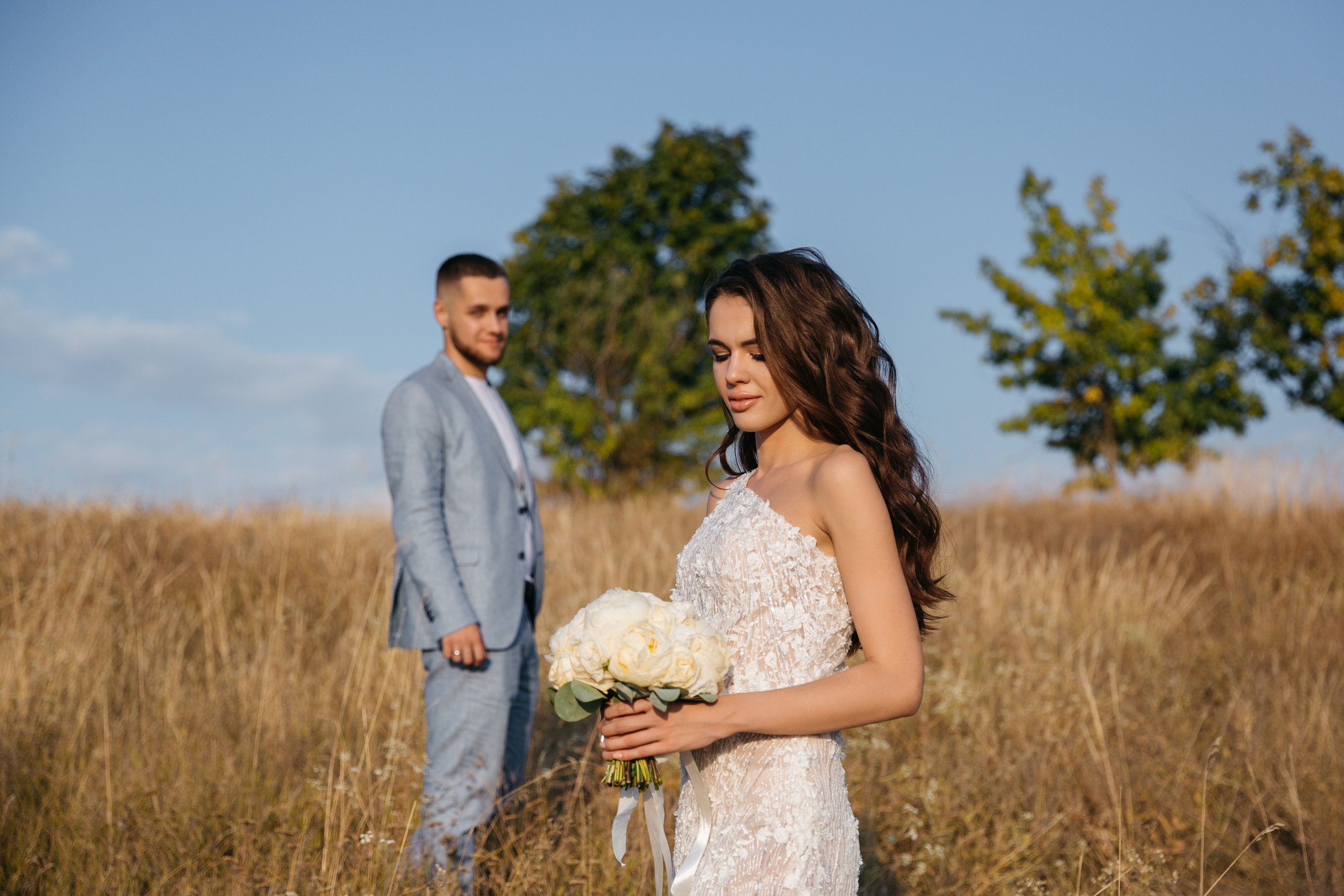 невеста с букетом в поле позирует и на нее смотрит влюбленно жених