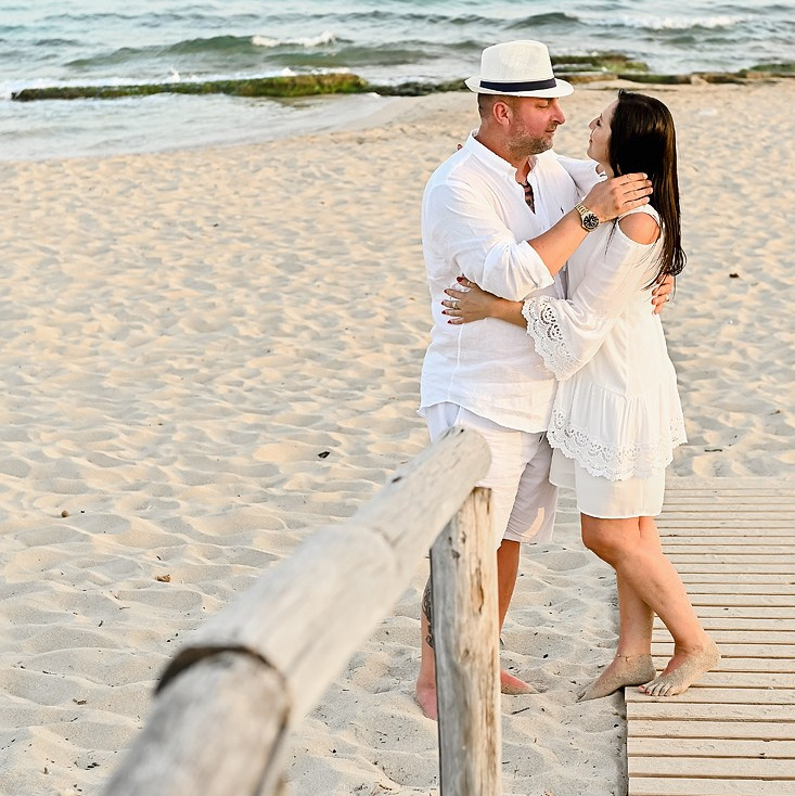A couple embraces on the beach at sunset, dressed in white, with the ocean and a wooden walkway in the background.