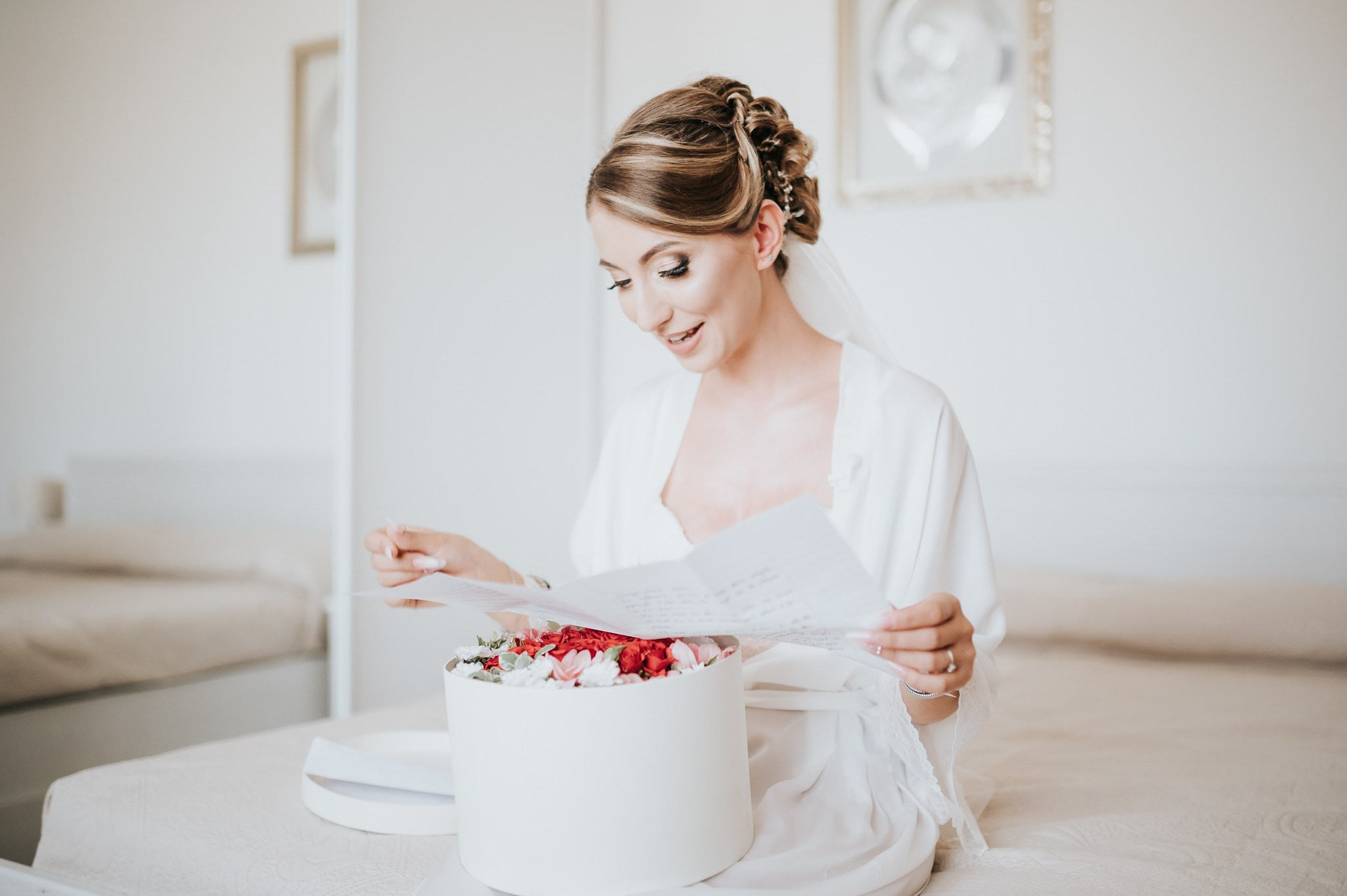 Sposa affascinante legge una lettera commovente, seduta accanto a una scatola elegante di fiori freschi, in una stanza raffinata che enfatizza la dolcezza del momento prima della cerimonia nuziale.