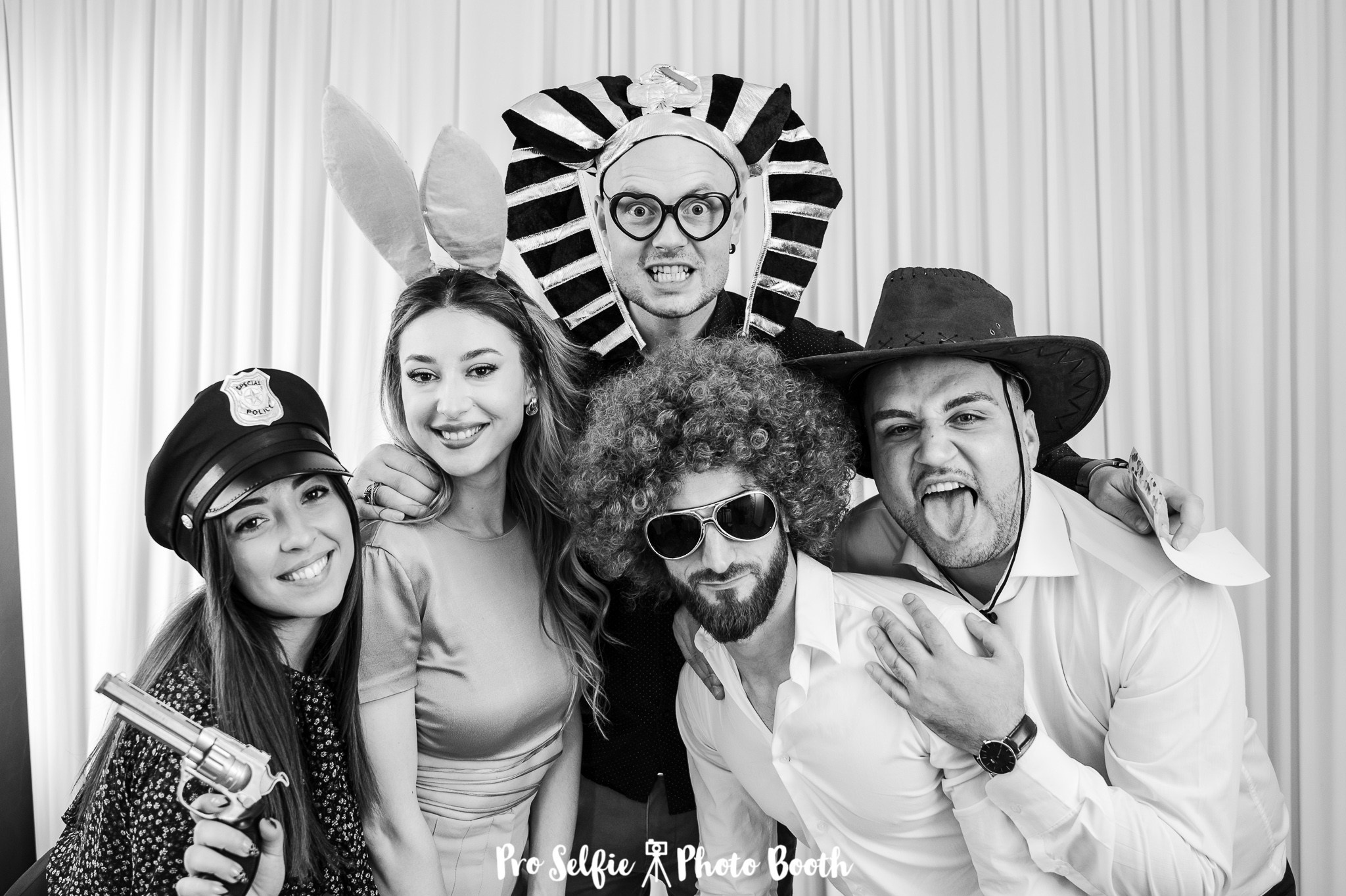 Gruppo di amici si diverte in un photo booth matrimoniale, indossando accessori divertenti e mostrando espressioni esuberanti, immortalati in una foto ricordo in bianco e nero piena di allegria e spirito festoso.