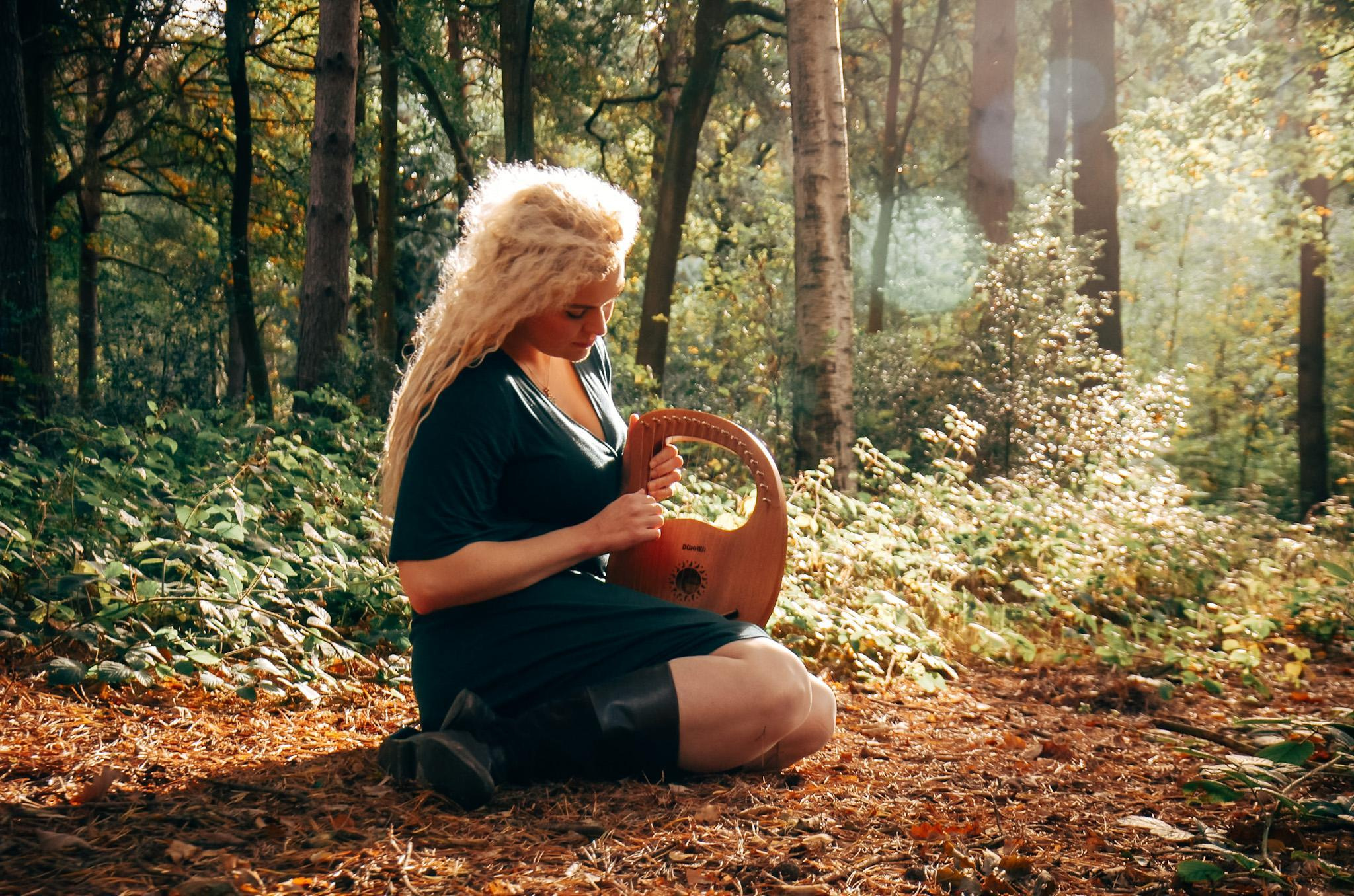 A beautiful blond girl playing a musical instrument in the woods in a green dress. Individual artistic photoshoot.  
