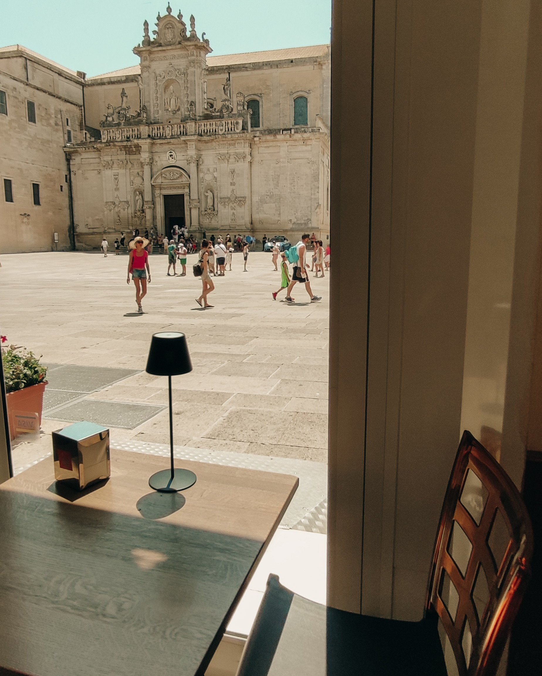 View of Lecce's cathedral square seen through the window of a cozy café. A hot midday, cool shade, and the slow rhythm of the city.