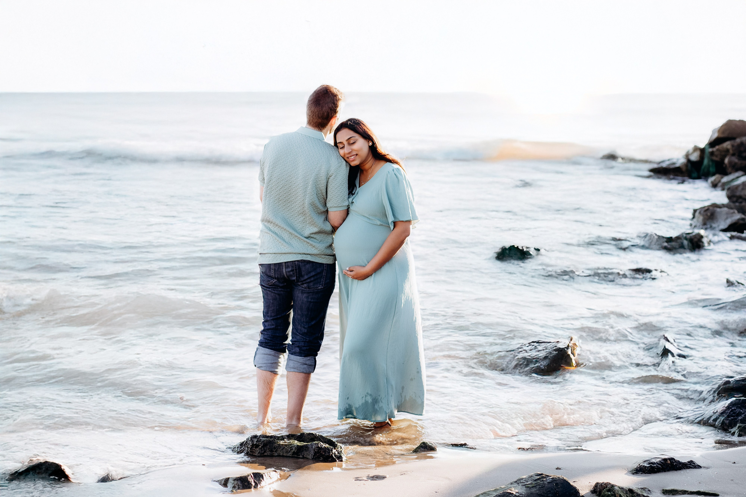 Romantic maternity photoshoot in Valencia, Spain, capturing an expecting couple standing together at the water’s edge on a Mediterranean beach, photographed in soft natural light with gentle waves and coastal rocks in the background, creating a peaceful and timeless pregnancy moment.