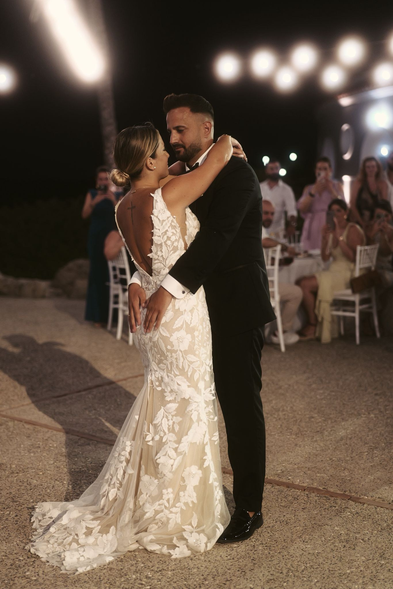 Romantischer erster Tanz eines Brautpaares bei einer Abendhochzeit in Kallithea Springs. Braut in floralem Spitzenbrautkleid, Bräutigam im schwarzen Anzug. Hochzeitsfeier mit Gästen und Lichterketten. Hochzeitstanz-Fotografie.