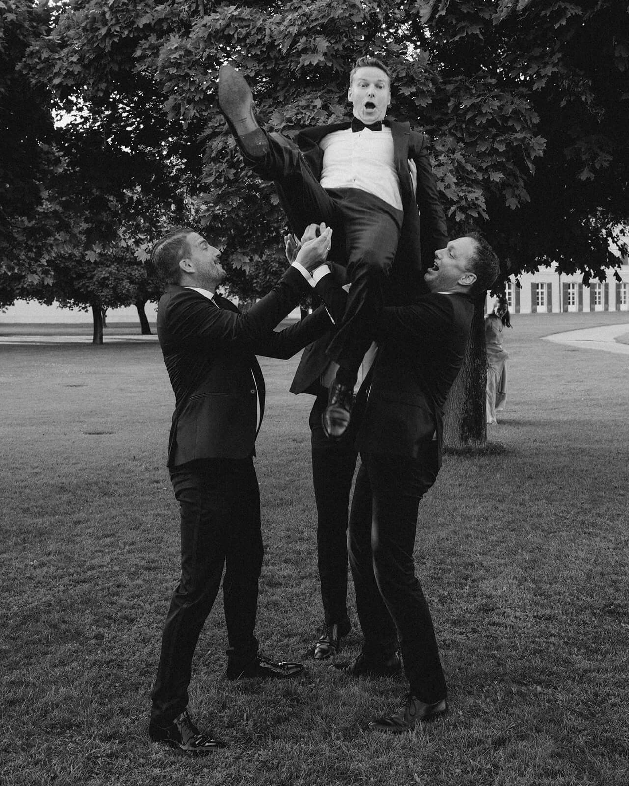 Two guests in black tuxedos hoist a wide-eyed groom into the air on the lawn of a grand estate: a candid black tie wedding moment