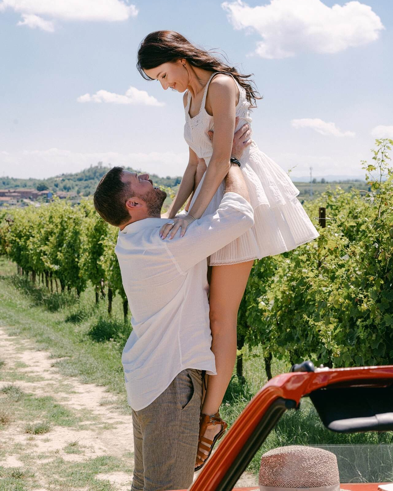 Жених поднимает невесту рядом с виноградниками Ланге и красным кабриолетом во время свадебной съемки в Пьемонте