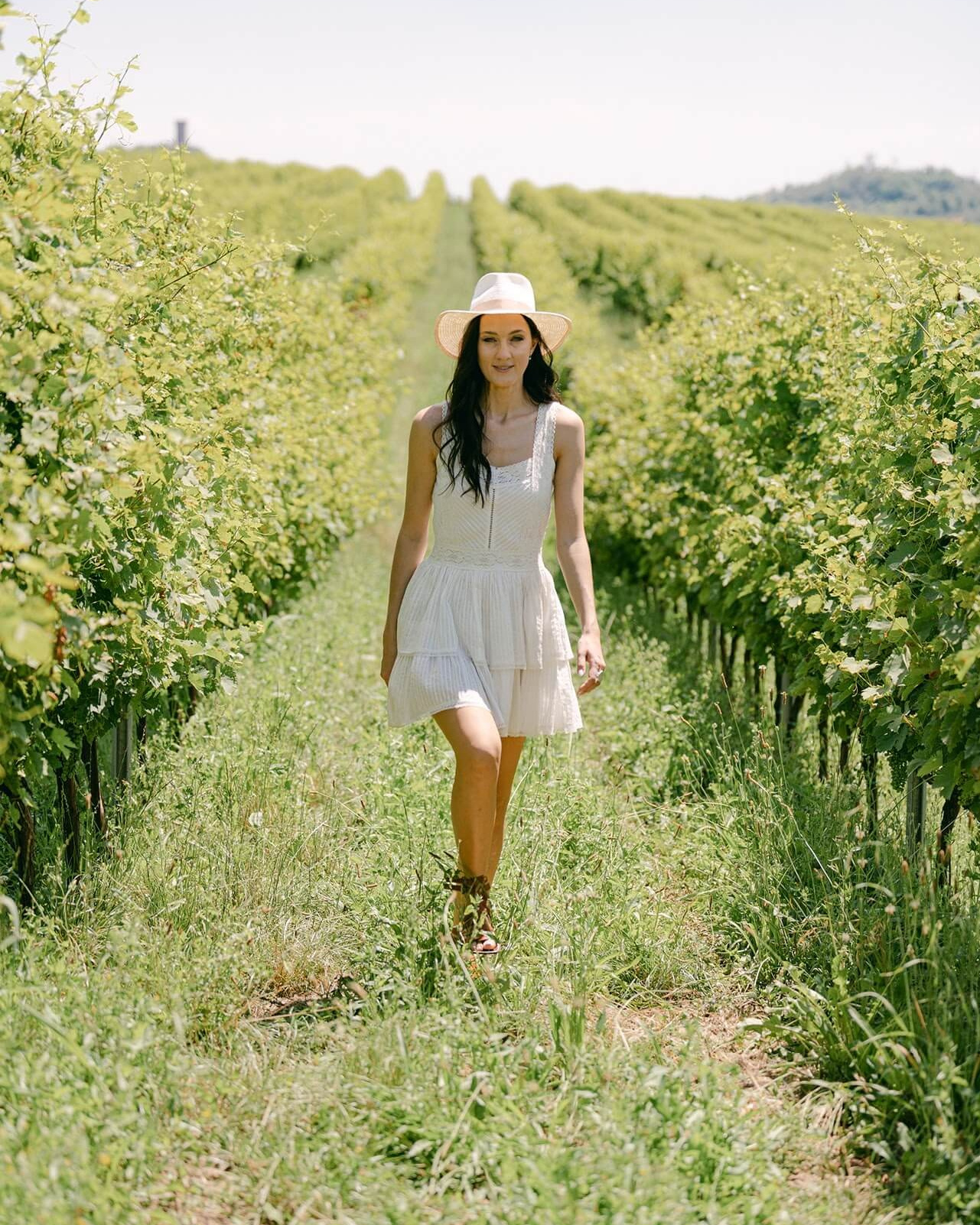 Невеста в коротком белом платье и шляпе идет между рядами виноградников Ланге во время свадебной съемки в Пьемонте