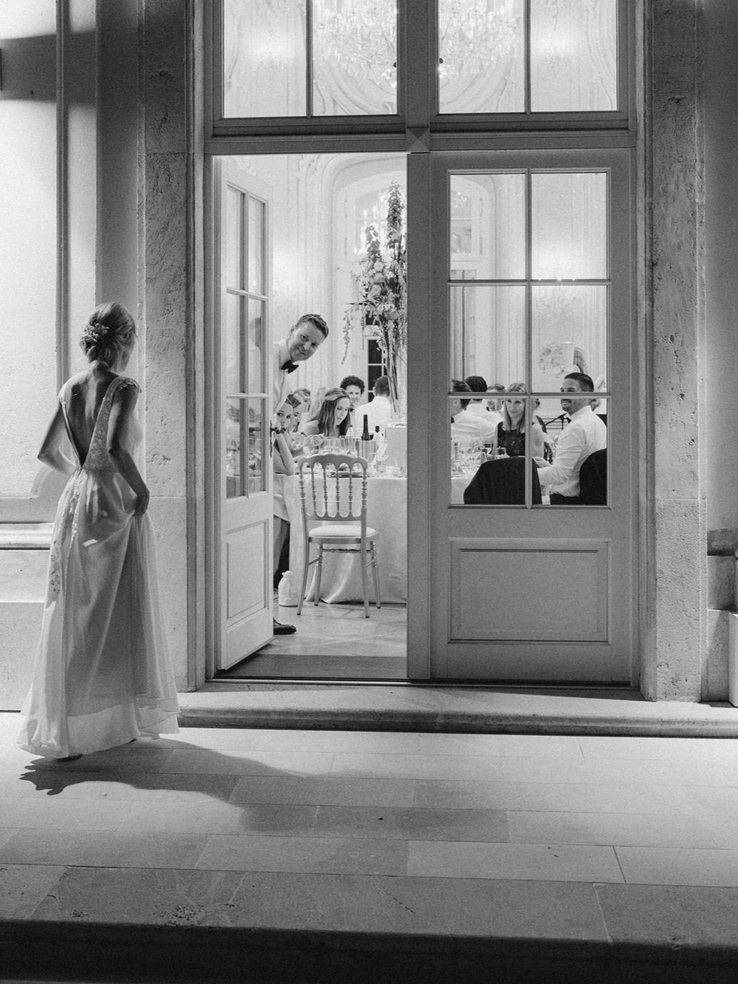 Bride at the threshold of open French doors, the groom glancing back as a chandelier-lit luxury reception room glows behind him