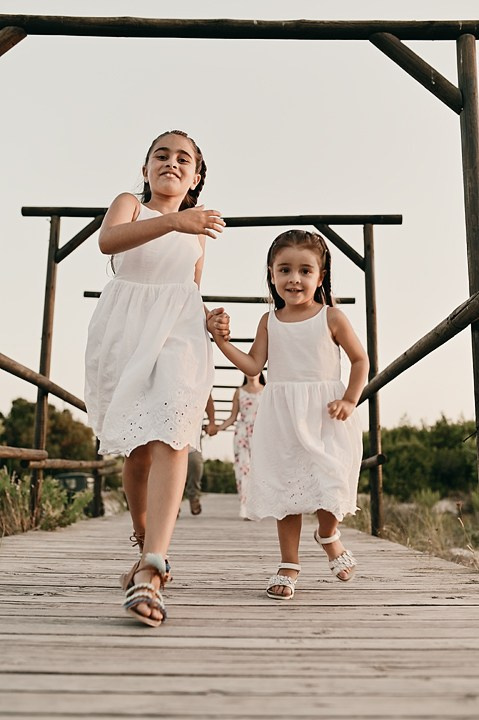 Two girls in white dresses are running along a wooden bridge with a summer landscape in the background.