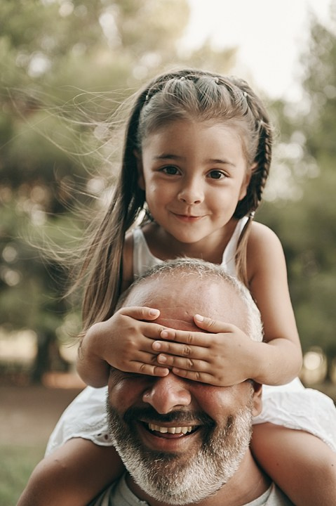 A sweet portrait of a little girl covering her father’s eyes while sitting on his shoulders, both laughing together.