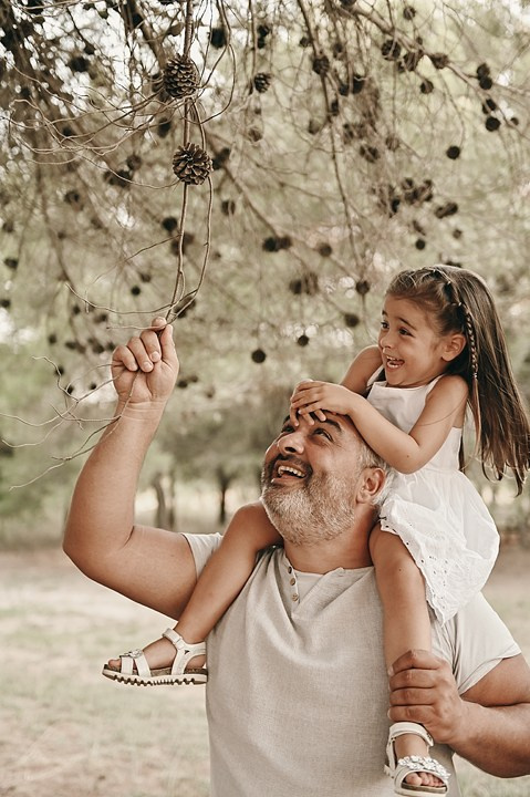 A joyful moment of a father carrying his granddaughter on his shoulders under a pine tree, both smiling as they play with the hanging cones.