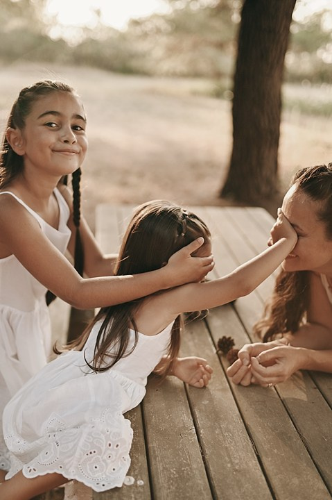 One girl gently covers her mom’s eyes while her sister gives a cheeky smile — a playful moment at a wooden table in the forest.