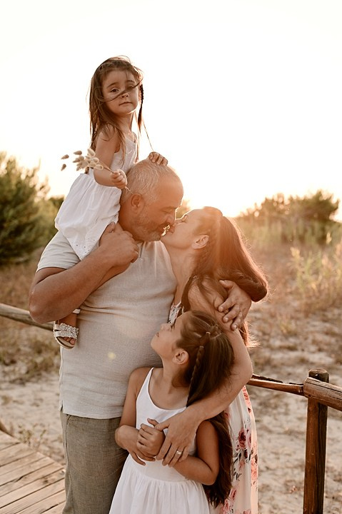 A family embraces at sunset — the father holds his daughter on his shoulders while the mother kisses his cheek and their younger girl hugs him.