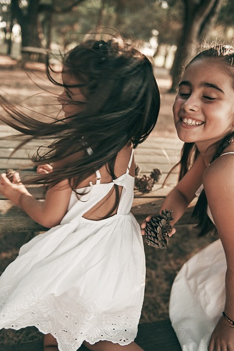Two sisters in white dresses playing with pinecones on a wooden bench, their hair flying in the wind as they laugh.