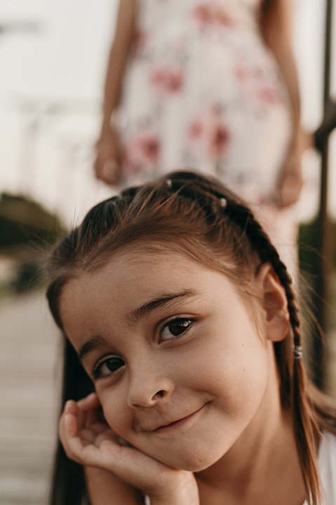 A girl in a white dress with braids smiles in a close-up, resting her cheek on her hand.
