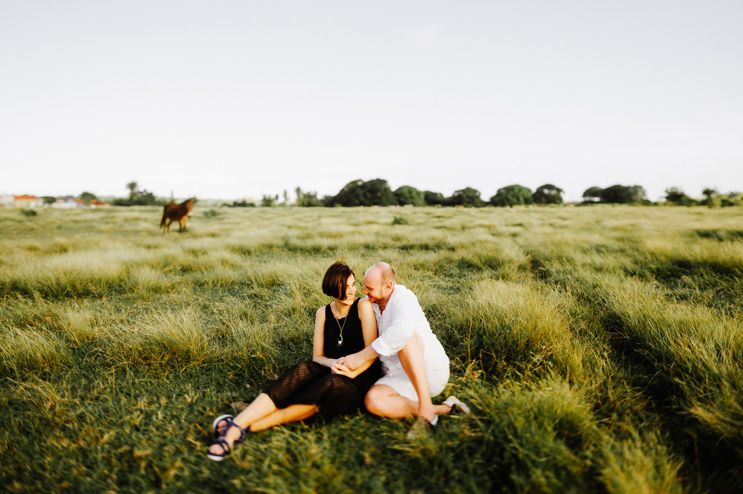 The newlyweds are sitting on the grass and hugging each other, the bride has a black dress and the groom is dressed in shorts and a white shirt.