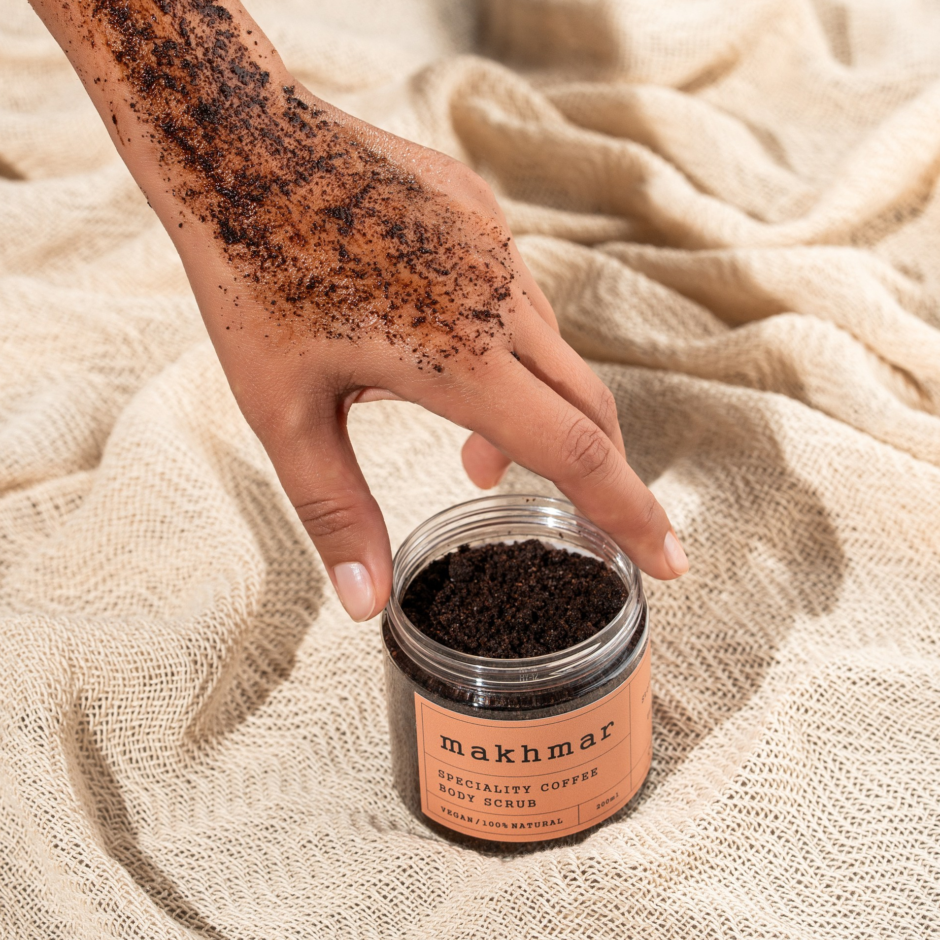 Əl üzərində Makhmar qəhvə skrabı tətbiq olunur, məhsul bankası bej teksturlu parçanın üzərində yerləşdirilib