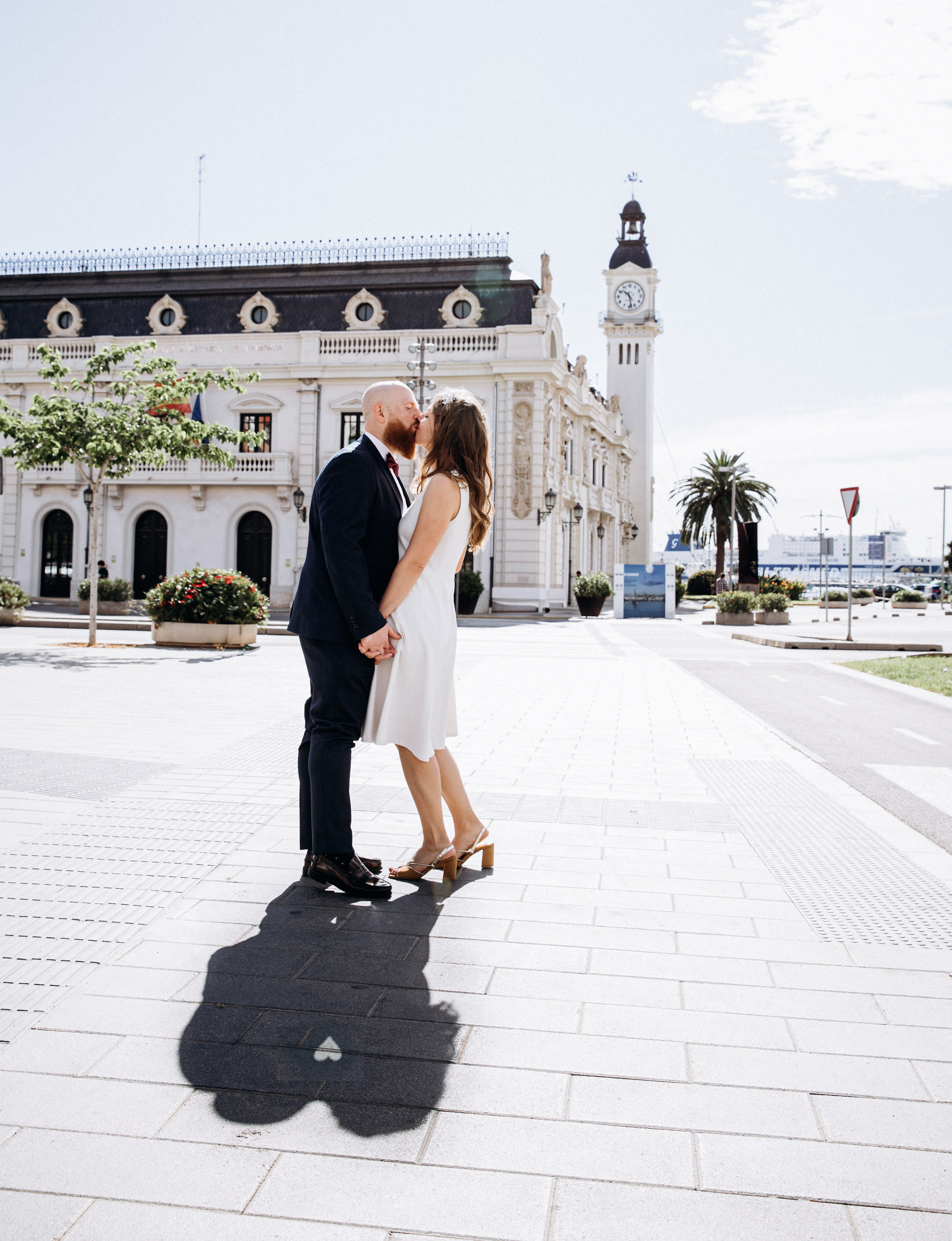 Couple kissing in a sunlit plaza, with an ornate historic building and clock tower behind them.