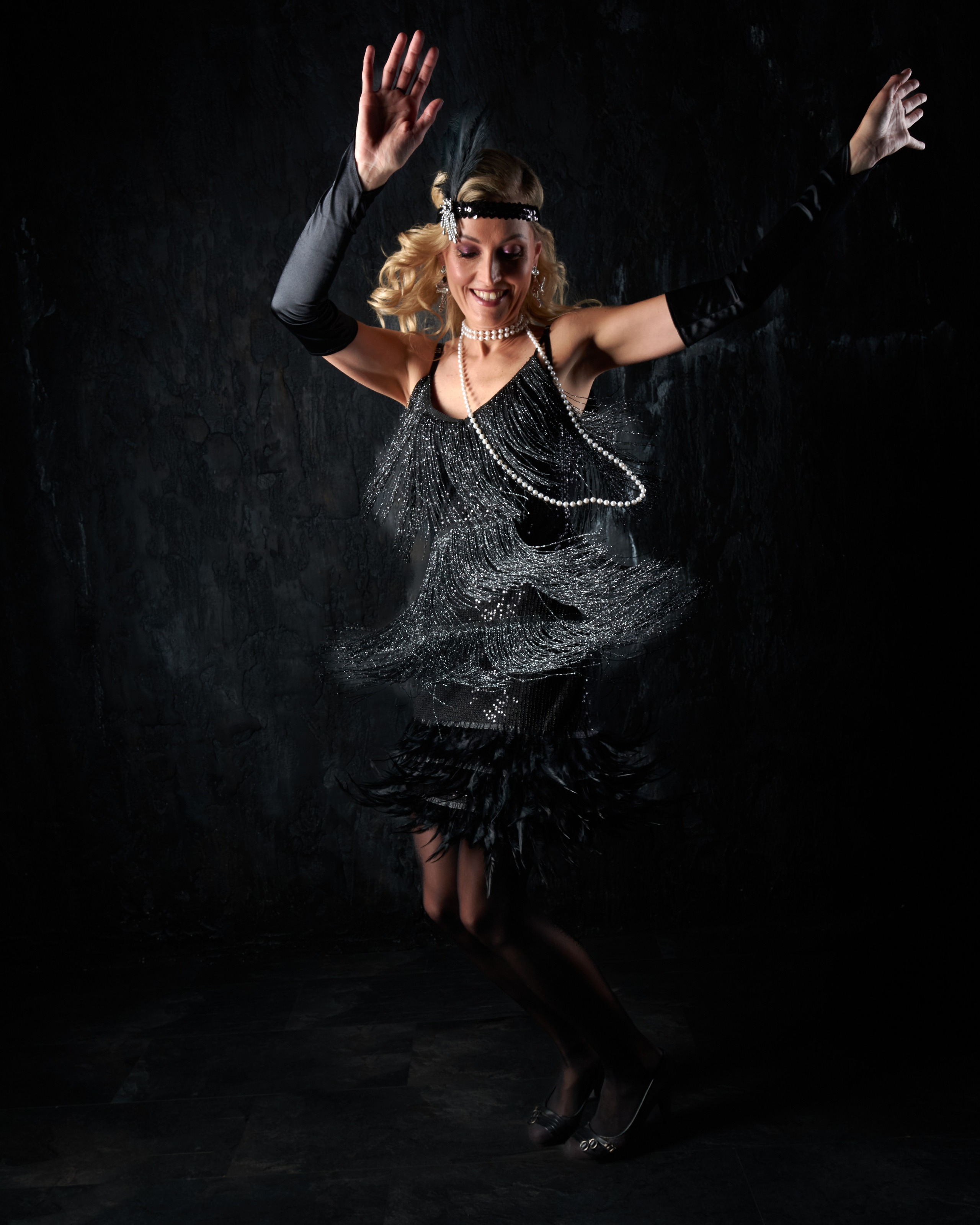 A woman as a flapper dances with raised arms and a smile, dress fringe caught in motion