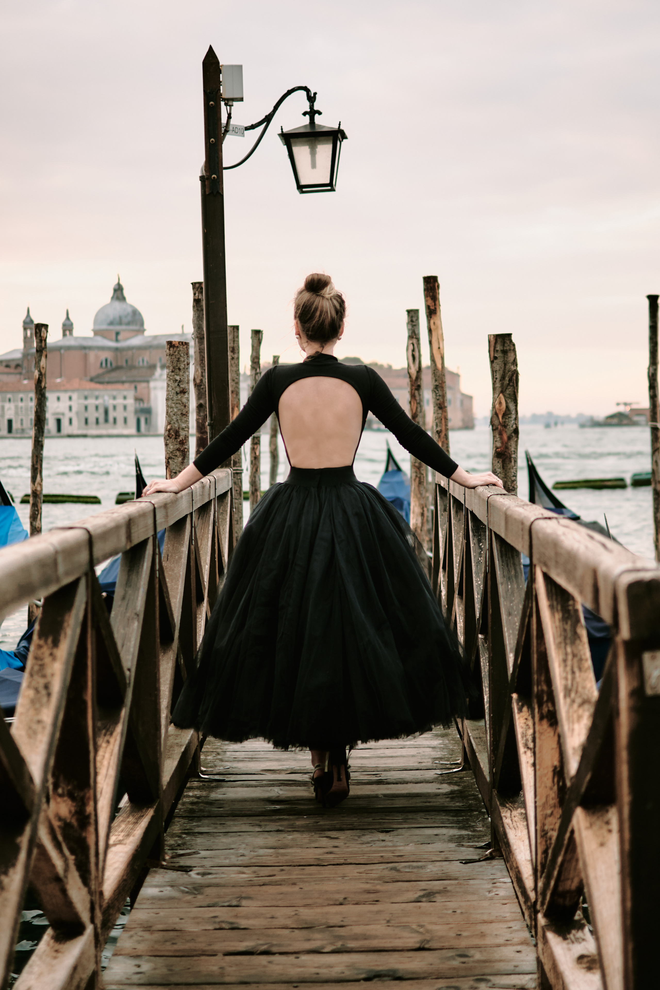 A young woman stands on San Marco Square in Venice, Italy. She wears a flowing, black dress that catches the gentle breeze blowing in from the sea. In the background, the iconic lagoon of St. Mark's square. The bustling square is filled with people and the sounds of music and laughter, creating a lively and romantic atmosphere. Fashion blogger shoot Venice. Portrait and Fashion Photographer in Venice, Italy. Instagram Photos with lifestyle photographer