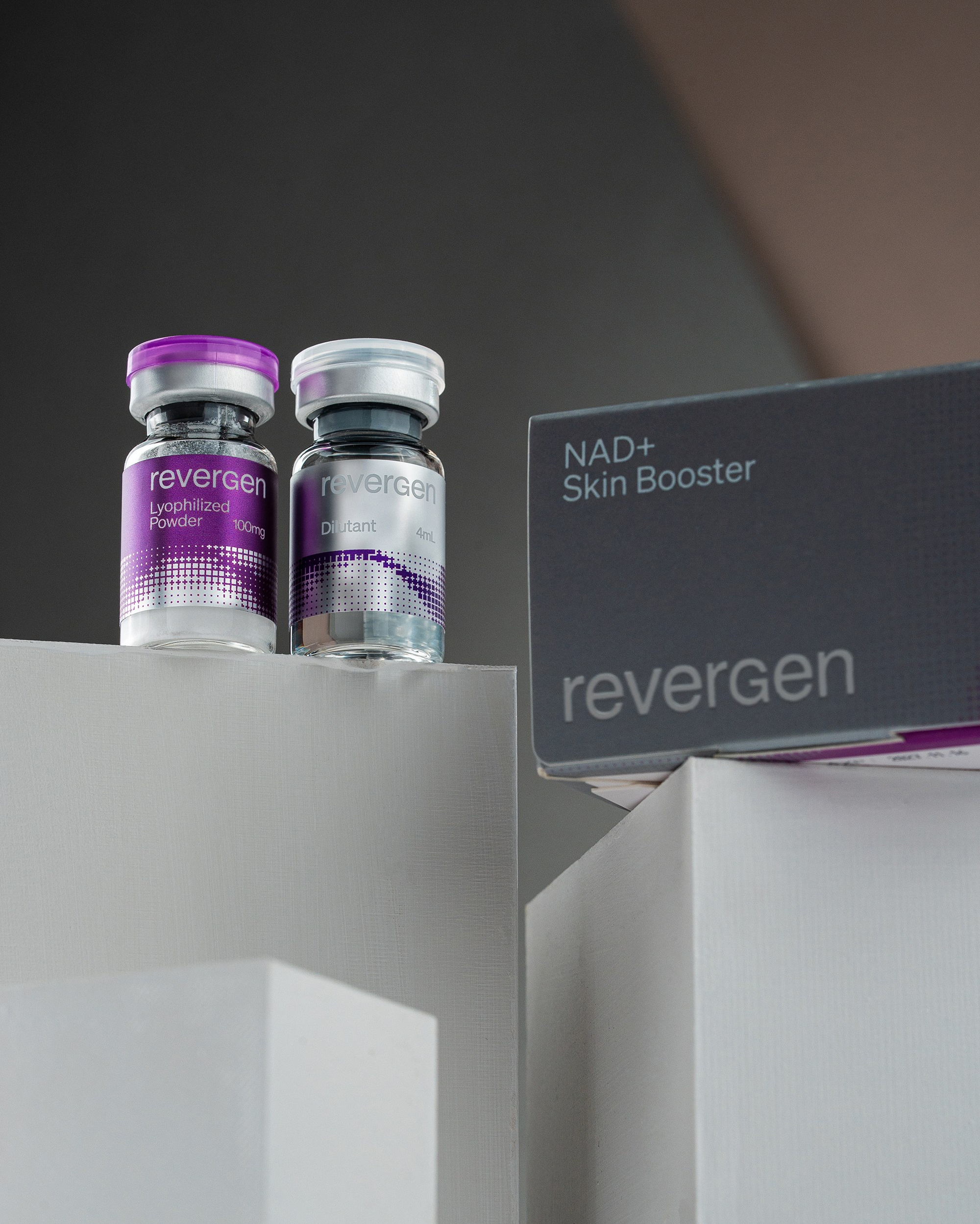 Revergen NAD+ Skin Booster kit displaying two glass vials (lyophilized powder and dilutant) resting on white blocks next to a grey branded product box.