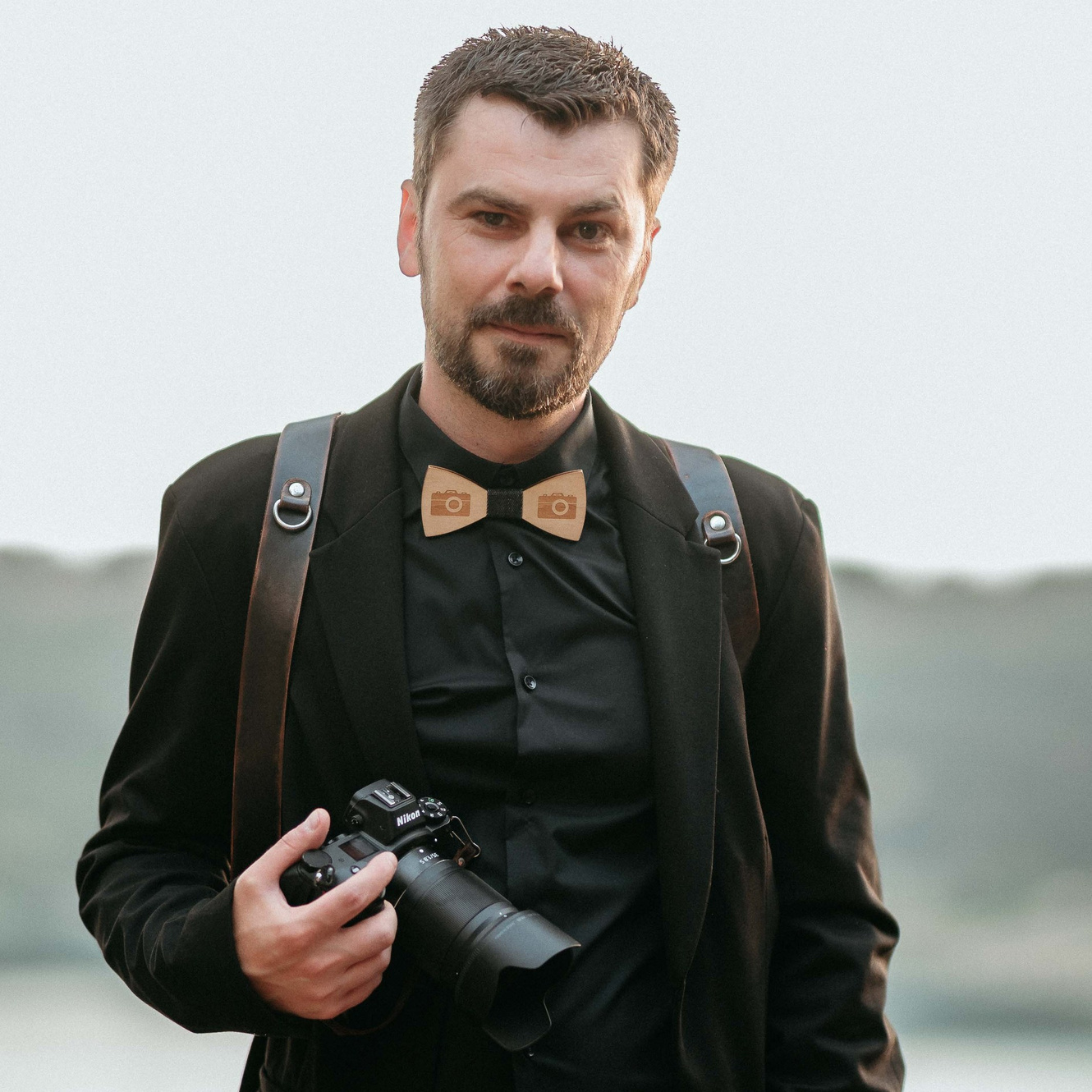Fotografo professionista con barba, vestito elegantemente in nero, che tiene una fotocamera reflex, sullo sfondo di un paesaggio naturale Lago di Castel Gandolfo