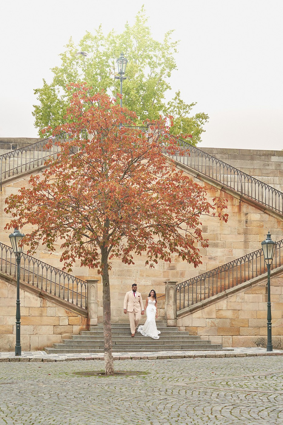 A tree vivid with Autumn colors stand near the Kampa steps as a groom and bride walk down.