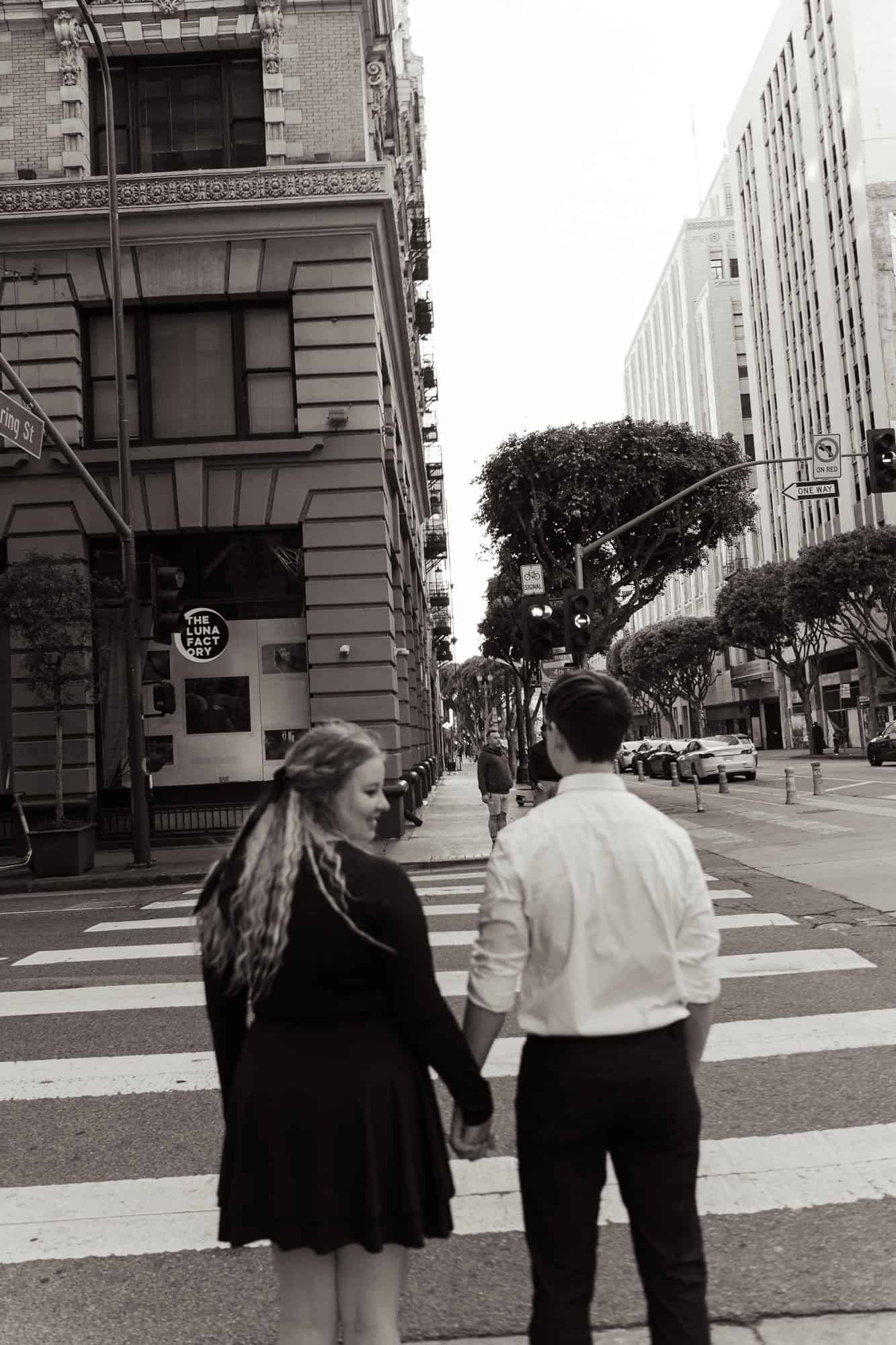 Cinematic engagement photo of couple crossing street with Los Angeles skyline in background