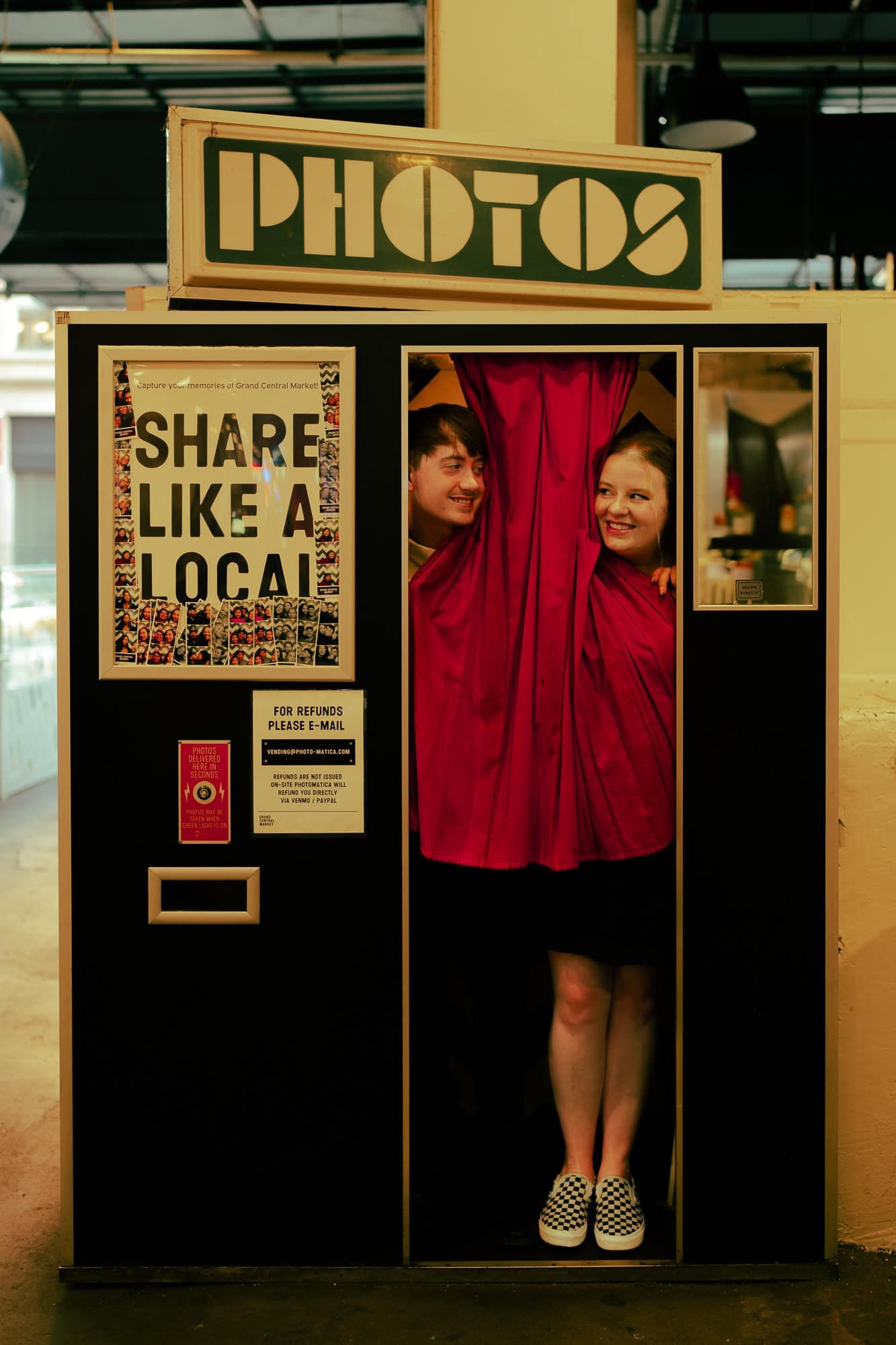 Romantic candid moment of an engaged couple peeking out of a retro photo booth with red curtains at Grand Central Market in Downtown Los Angeles, laughing and surrounded by vintage signage, perfect for an LA engagement session with a nostalgic twist
