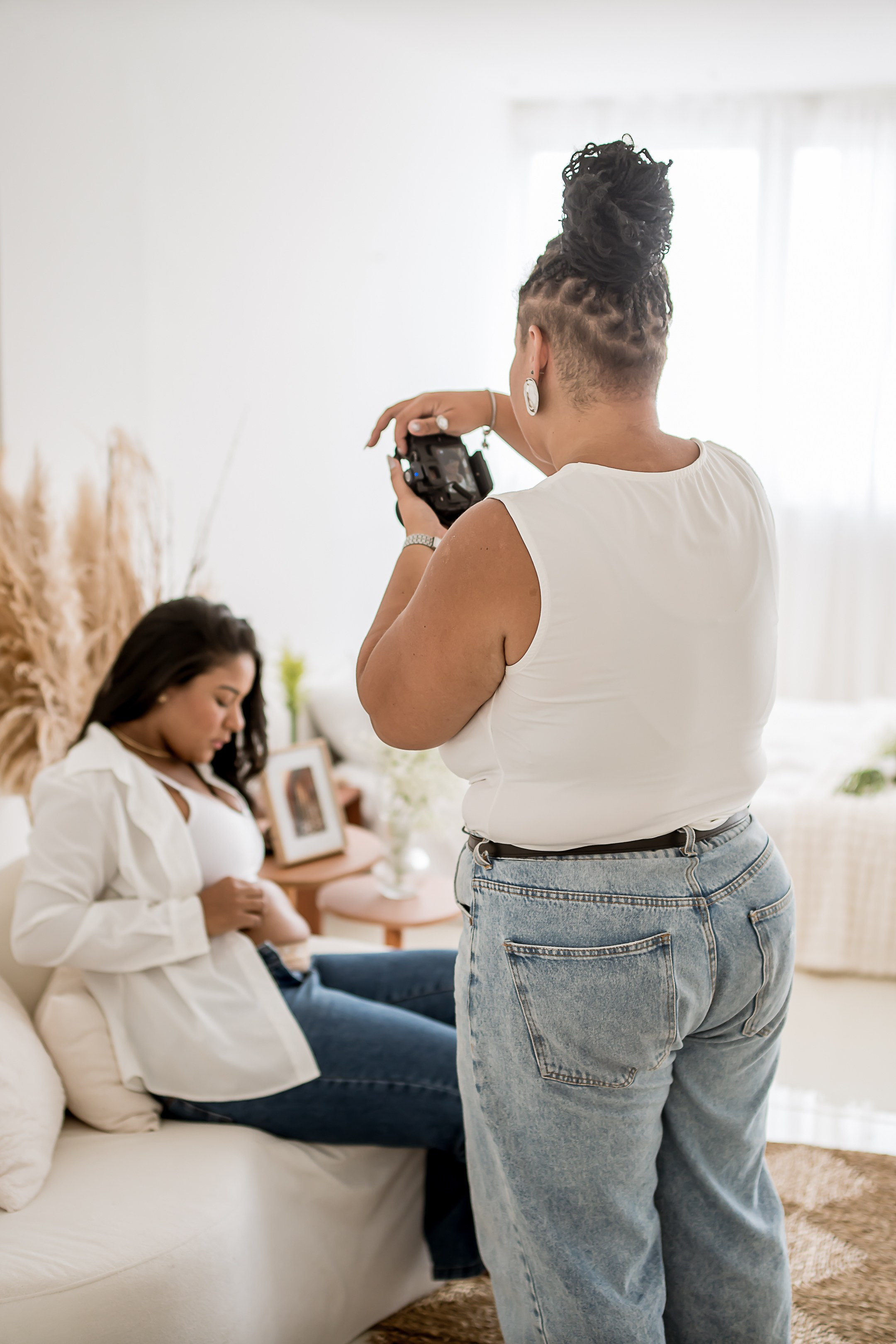 Fotografa de blusa branca, calça jeans fotografando gestante de jeans e blusa branca sentada em um sofá em estudio de luz natural. 