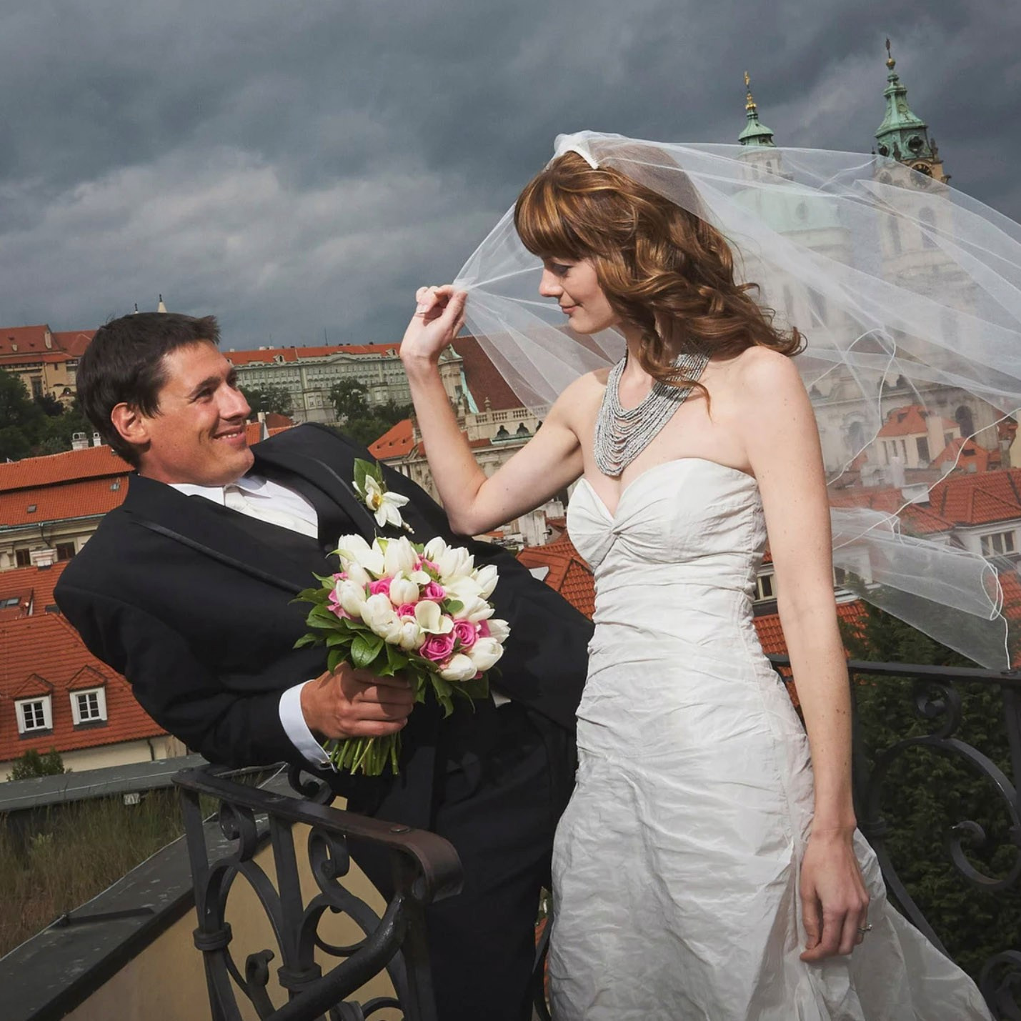 Red-haired bride holding her flowing veil aloft smiles seductively to her bouquet holding groom as he leans back against a railing in appreciation at the Vrtba Garden in Prague. 