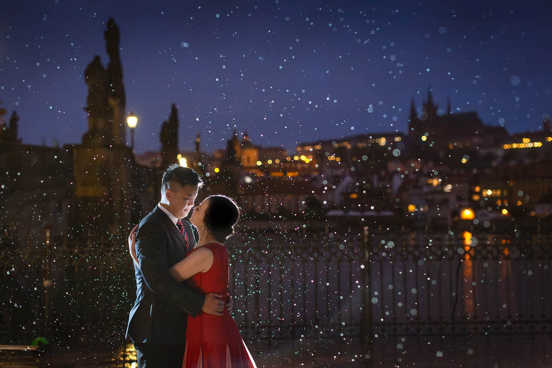 A couple embraces in the heavy rain near the Gothic tower leading to the Charles Bridge as the night skyline of Prague and the castle dominate the background.