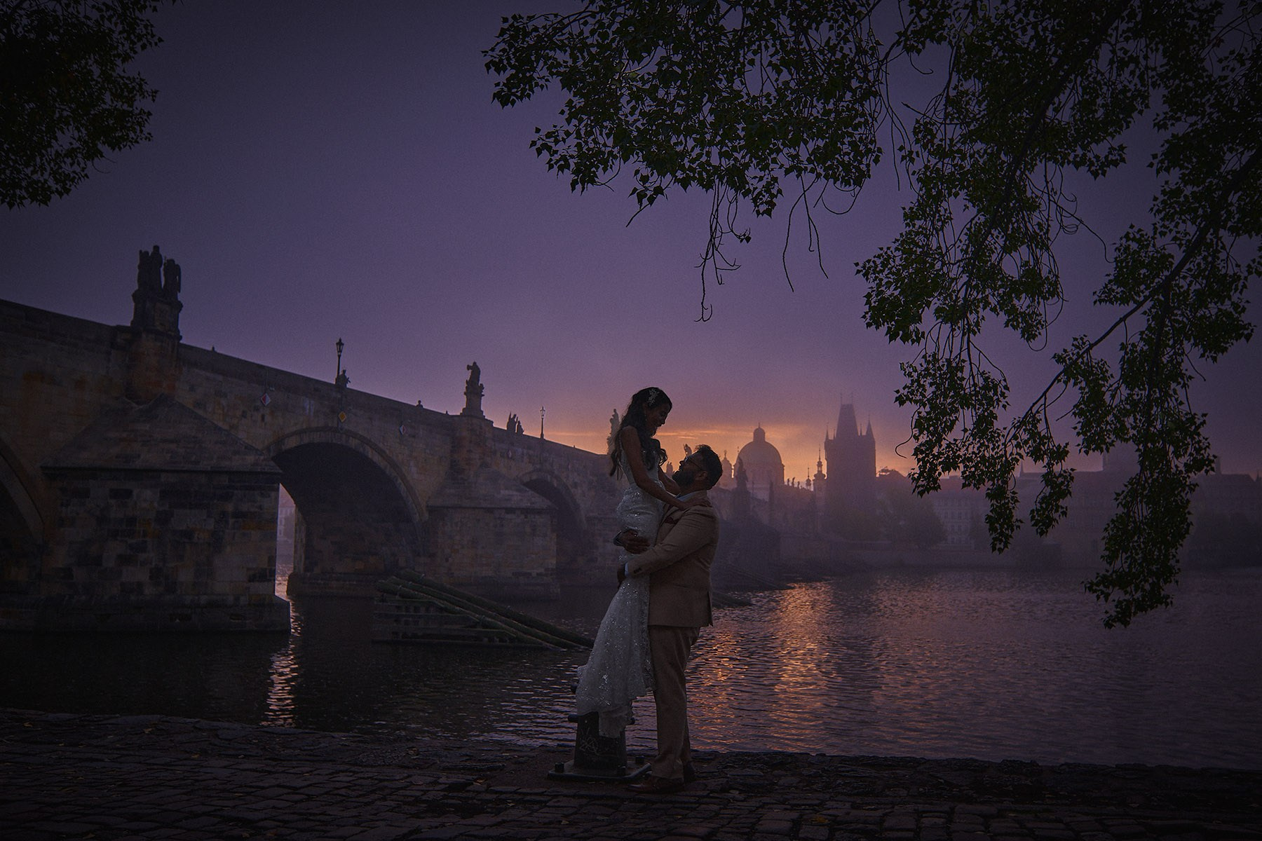 Bride and groom near the riverside as Charles Bridge is engulfed in fog as sky flares above