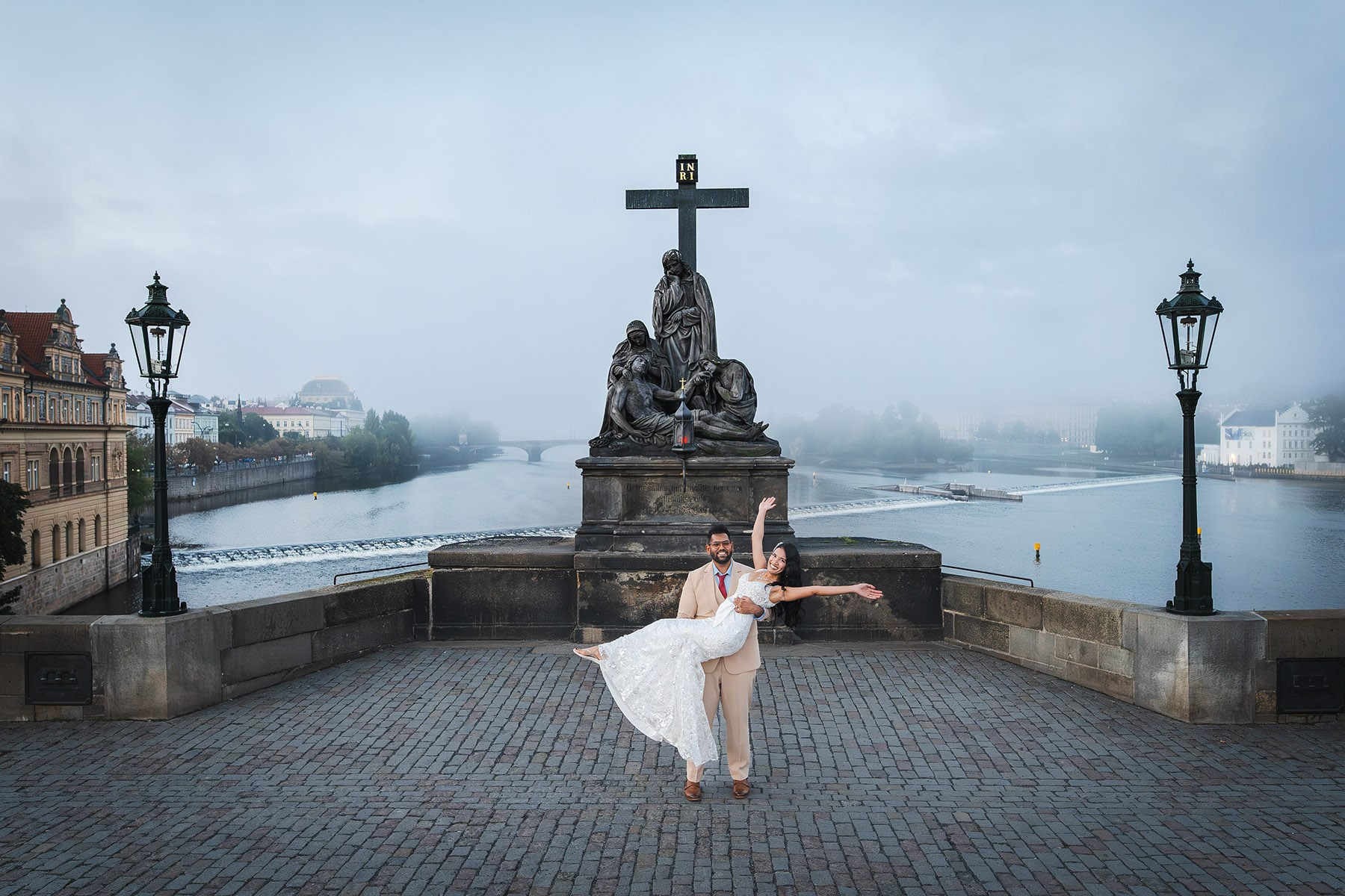 Groom carries his bride as she throws her arms up in excitement under the Pieta statue atop the Charles Bridge