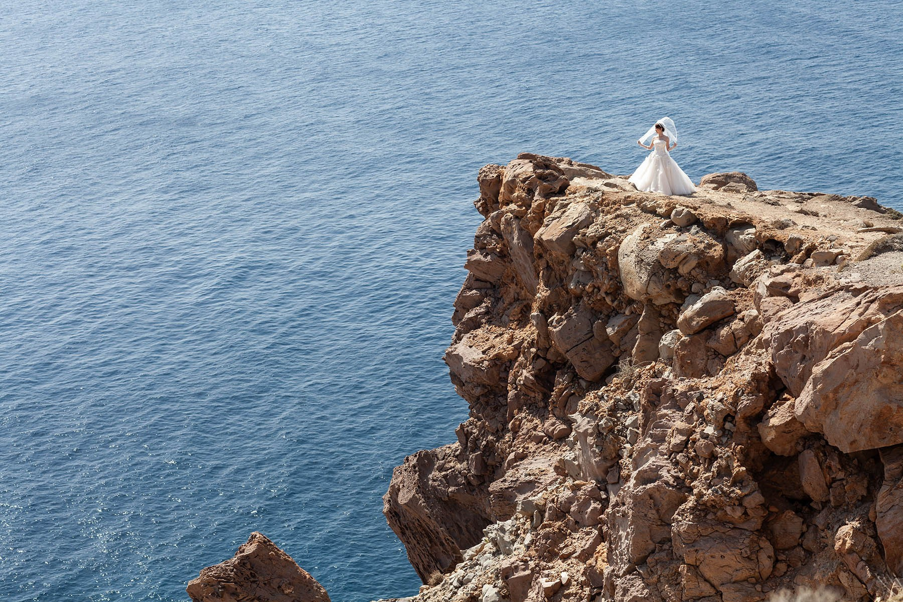 Bride holding her veil on the cliffs with panoramic views of the Aegean Sea in Santorini.