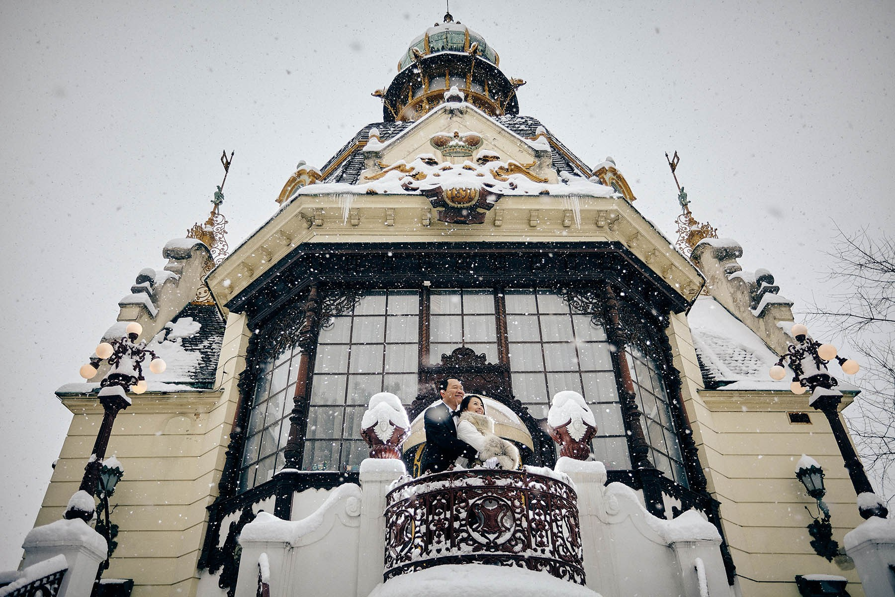 Couple on ornate snowy balcony at Hanavský Pavilion during heavy snowfall, Prague winter couples photography