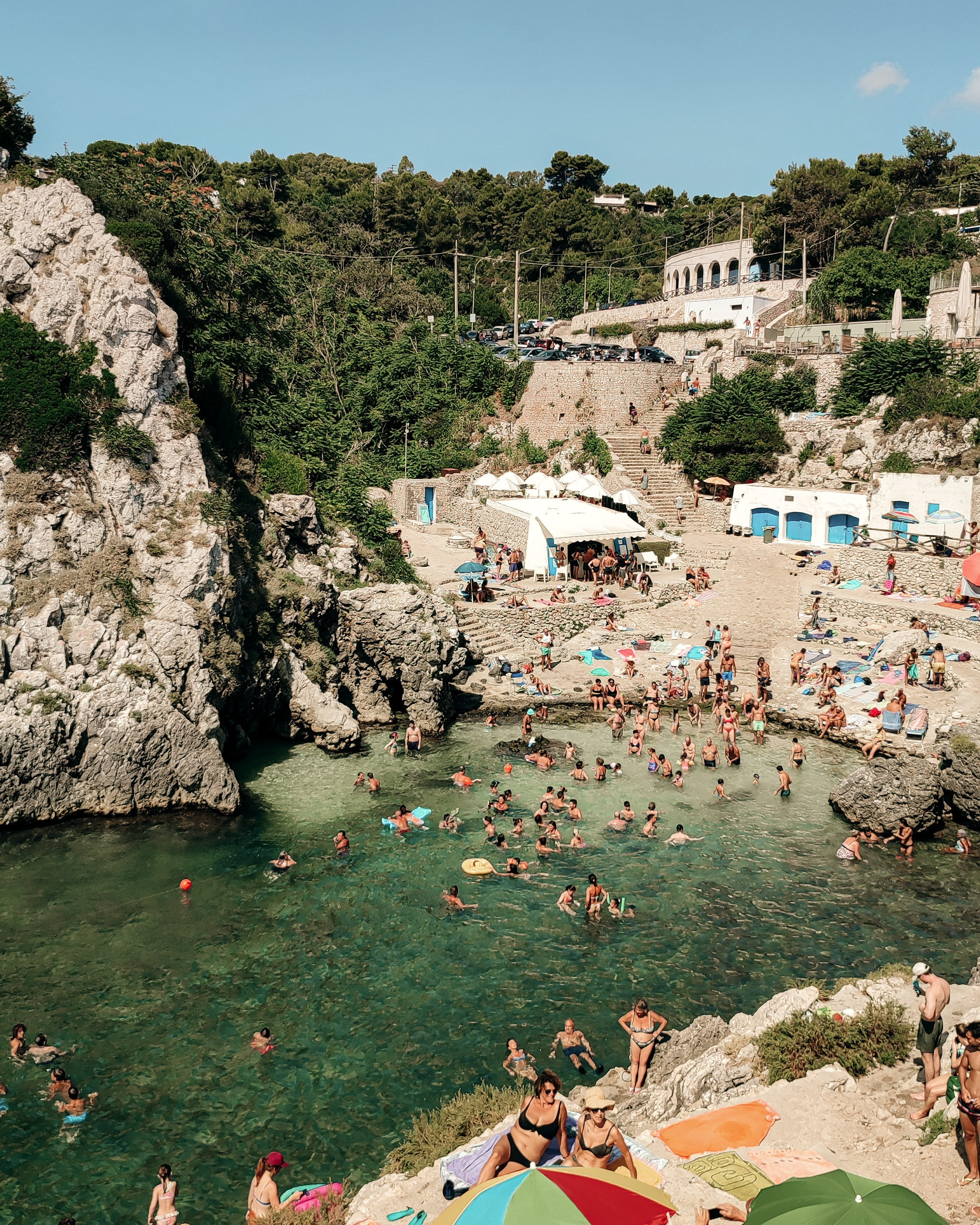 The scenic cove of Acquaviva with crystal-clear water and rocky surroundings. Locals and tourists swim and sunbathe on the rocks under colorful umbrellas.
