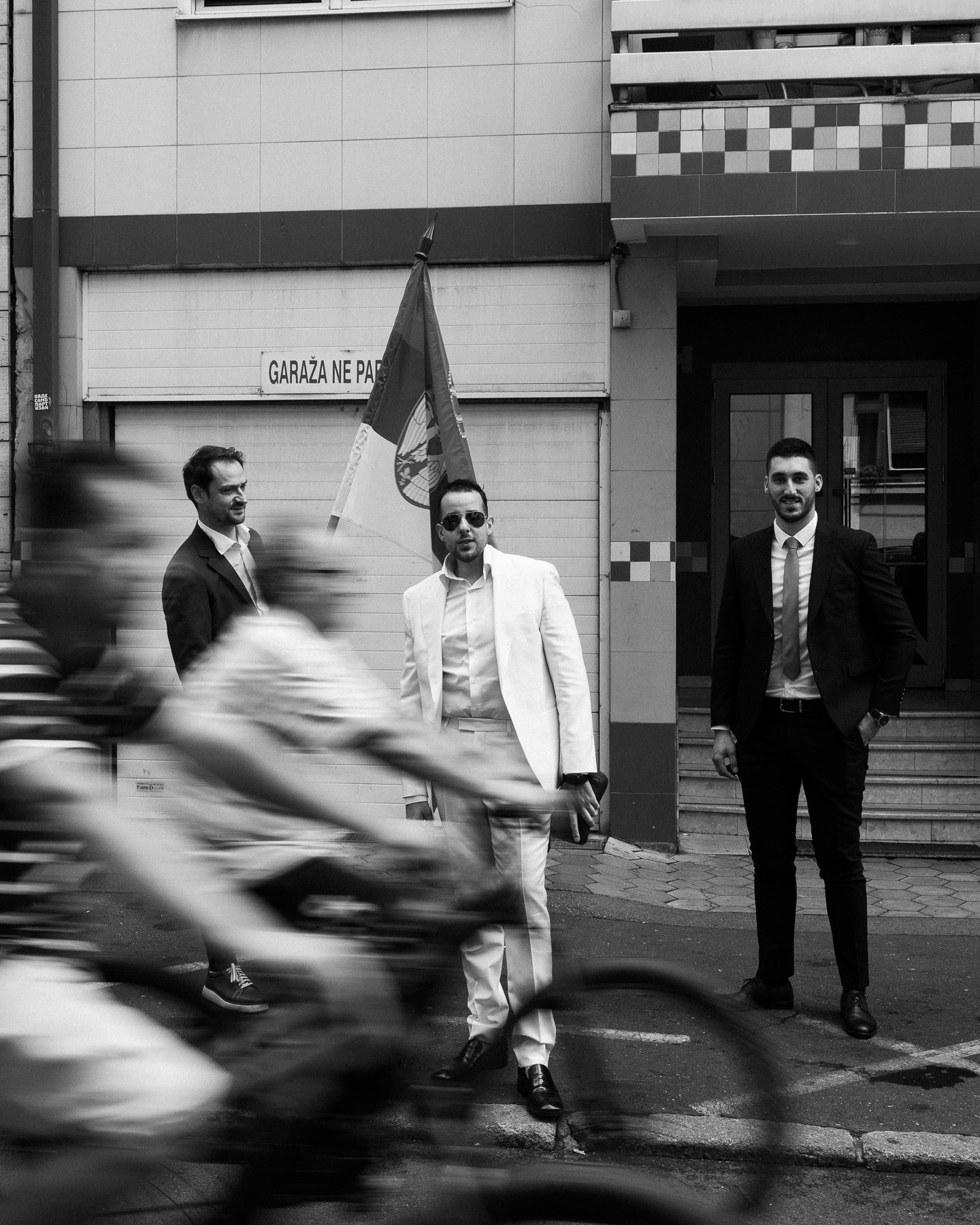 Crno-bela ulična fotografija trojice muškaraca u odelima na gradskom trotoaru sa zastavom, sa zamućenim biciklistima u prednjem planu; urbana scena.