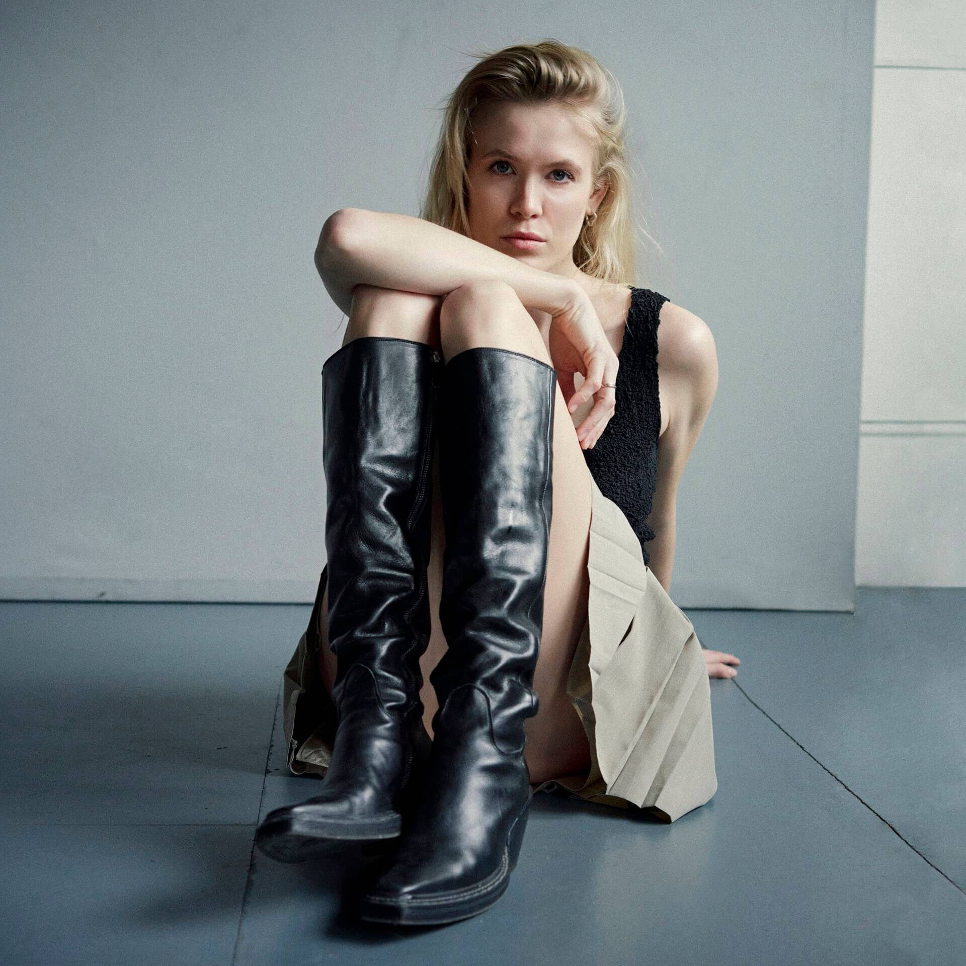 Blonde woman in black top sitting with tall black knee high boots fashion female portrait in Munich photostudio 